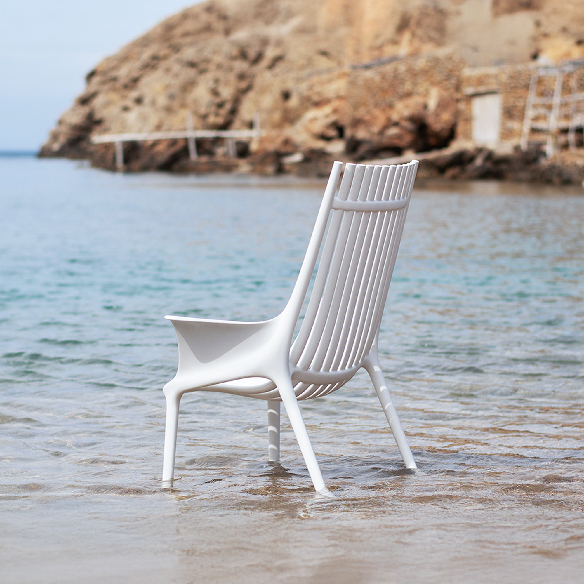 A modern white slatted chair stands in shallow, clear blue-green ocean water with a rocky cliff and rustic structures in the background.