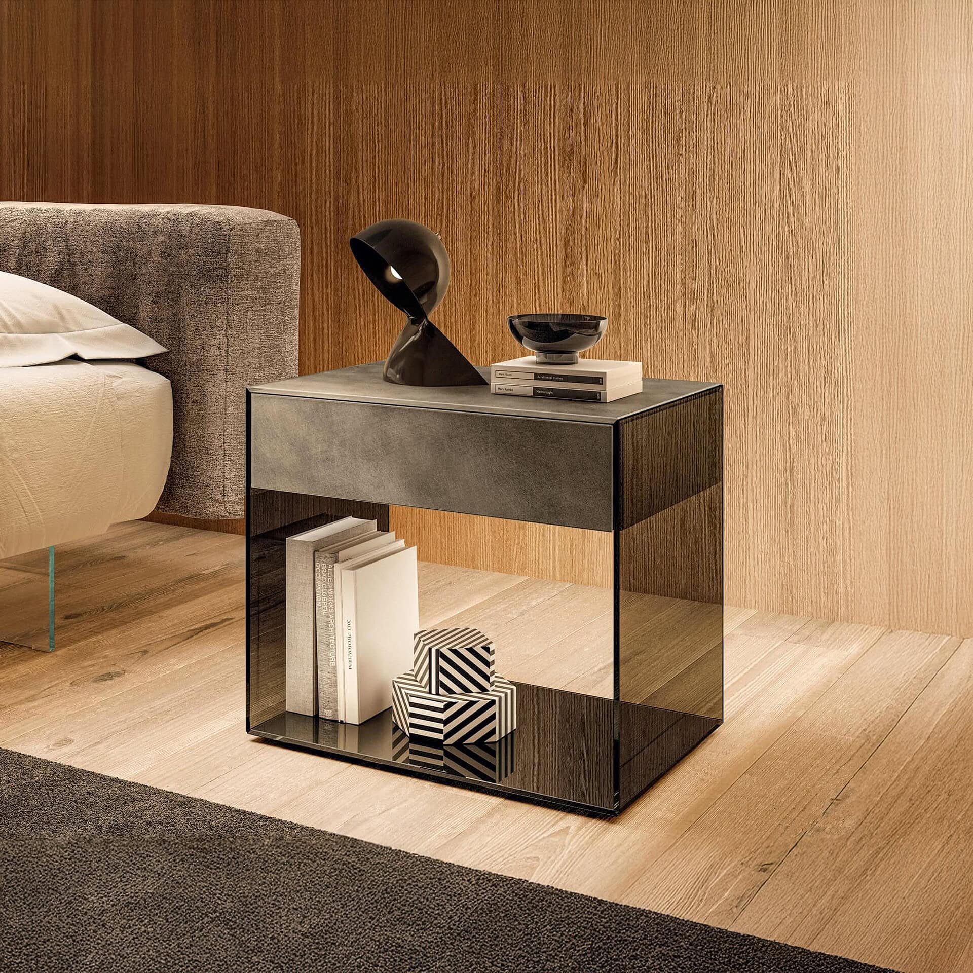 Lago UpGlass Bedside Table in black polished XGlass finish, displayed against a wood-paneled wall with decorative accessories.
