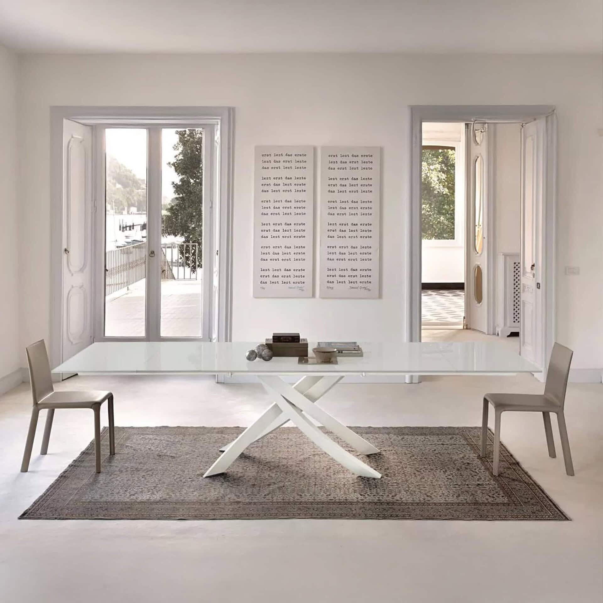 Artistico extendable dining table with a sleek glass top and sculptural crossed metal base, styled in a bright contemporary dining room.