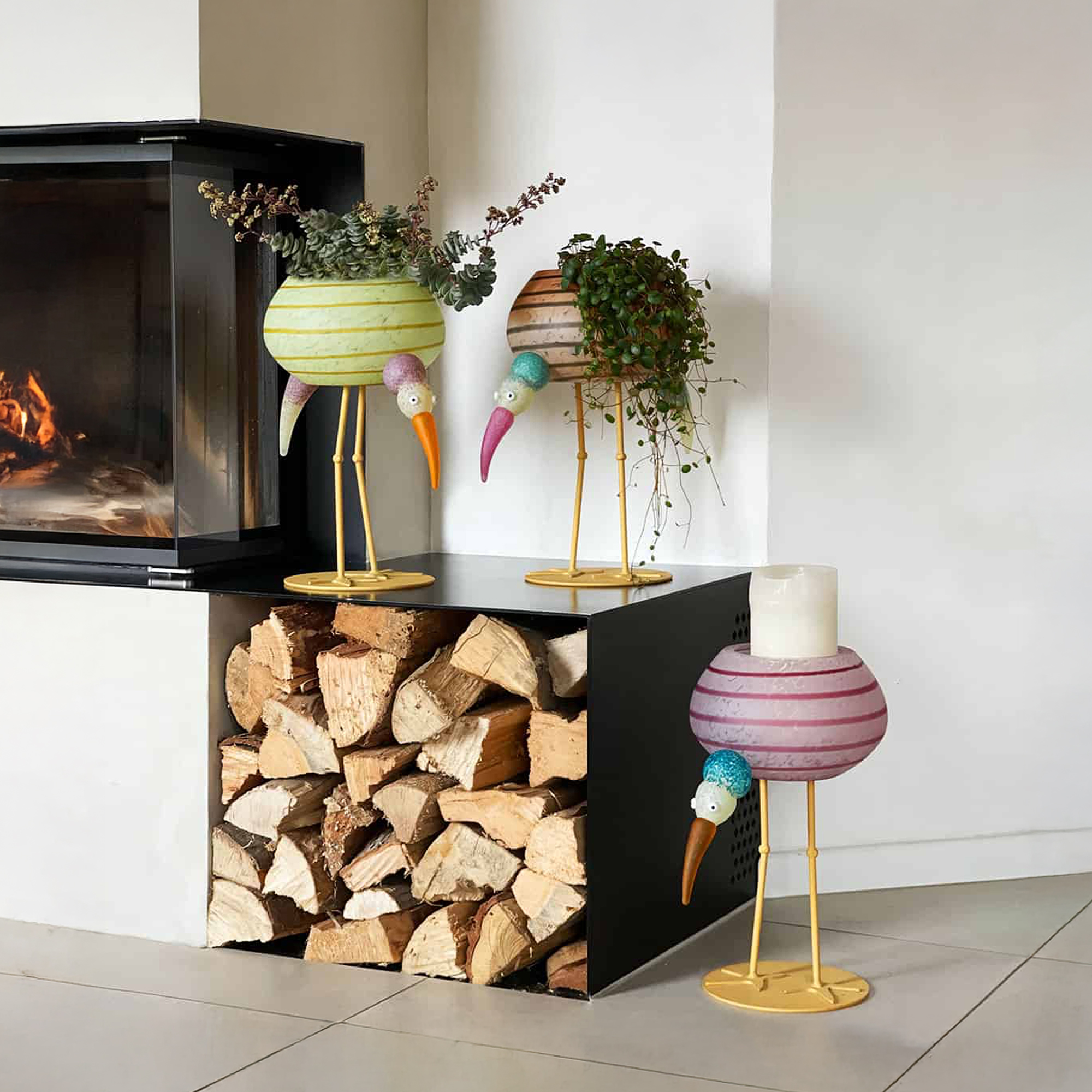 Borowski Ibis Glass Vessel in rose color displayed as a planter by a fireplace with wood storage.