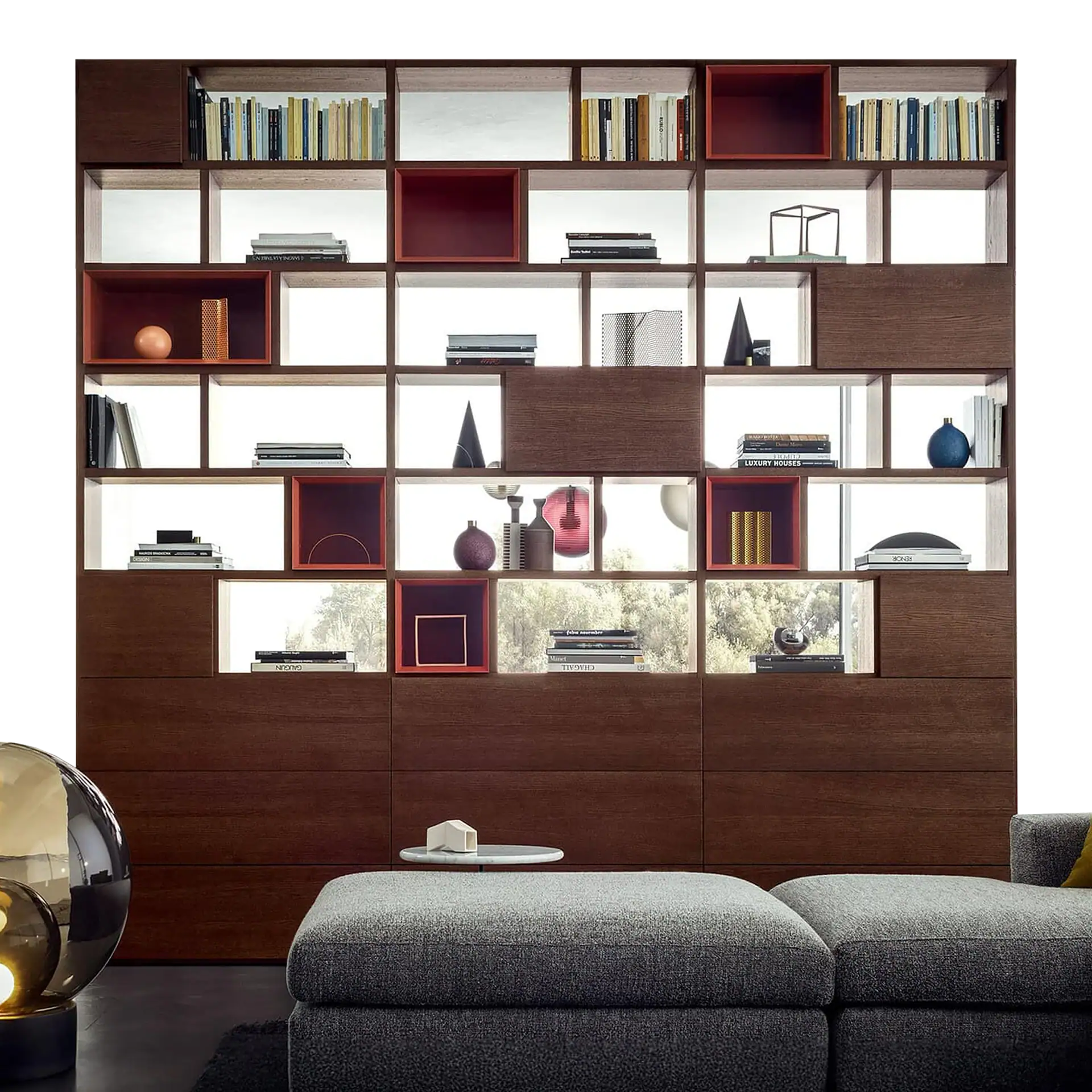 A modern dark wood shelving unit with illuminated compartments filled with books and decorative objects, including vases and sculptures. A grey upholstered ottoman and a spherical lamp are in the foreground.