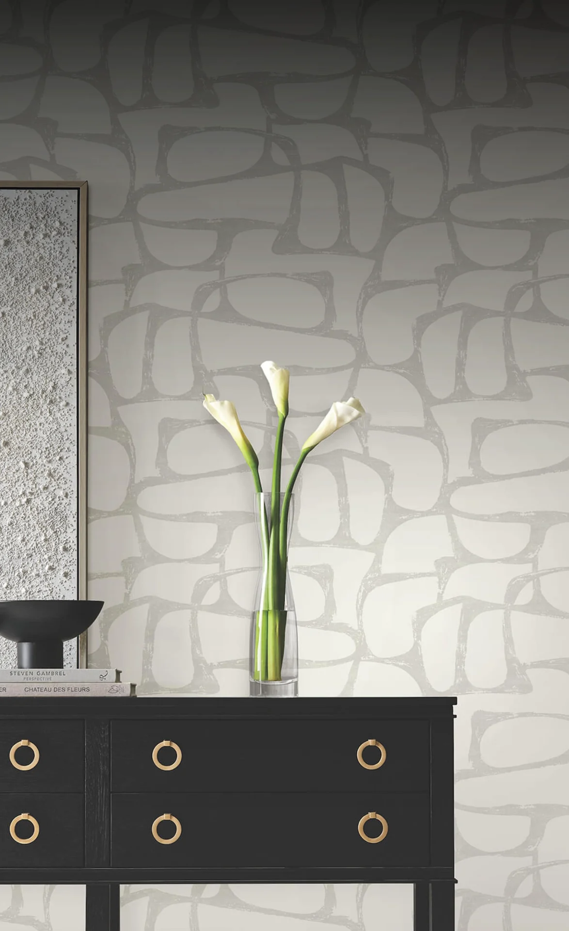A black console table with gold ring pulls is placed against a light gray wallpaper featuring an abstract, organic pattern. On the table, a textured white artwork, a black bowl, two books, and a clear vase with three white calla lilies are arranged.