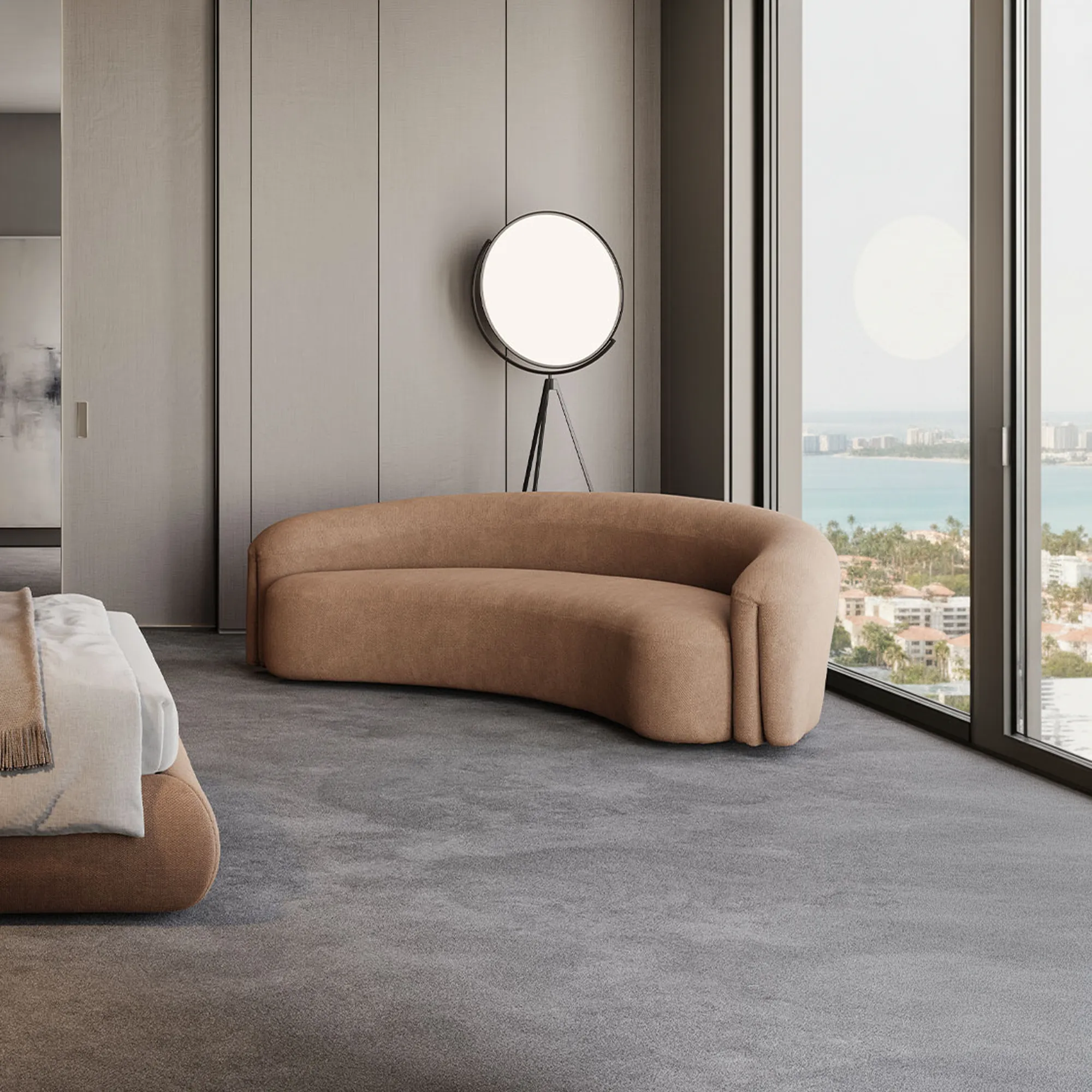 A modern living room with floor-to-ceiling windows showing a city view. Two curved taupe sofas and a round ottoman sit on a grey rug. A sculptural lamp and abstract art are also visible.