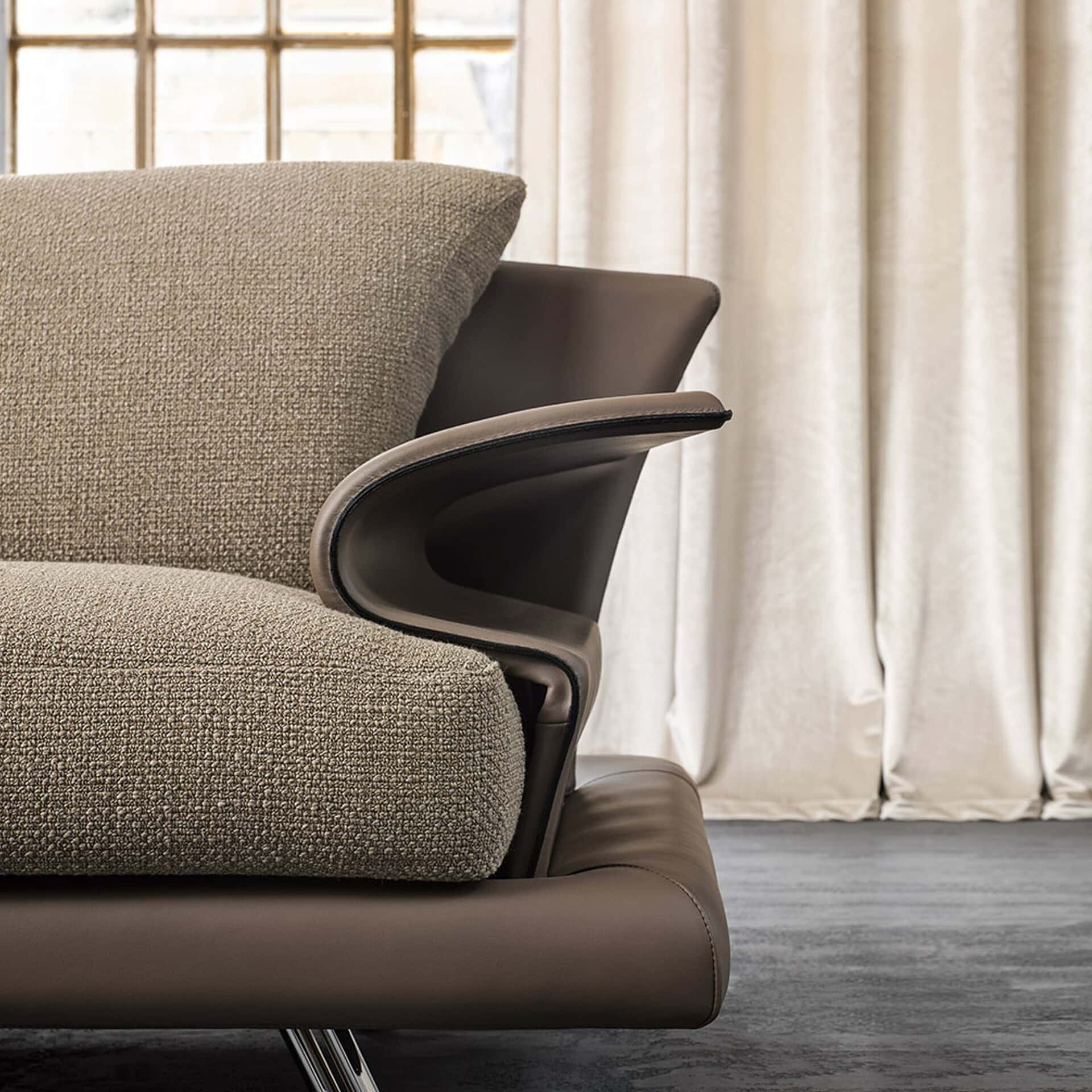 Close-up of a modern sofa with a textured light beige fabric cushion and a curved dark taupe leather armrest, set against a blurred background of cream curtains and a window.