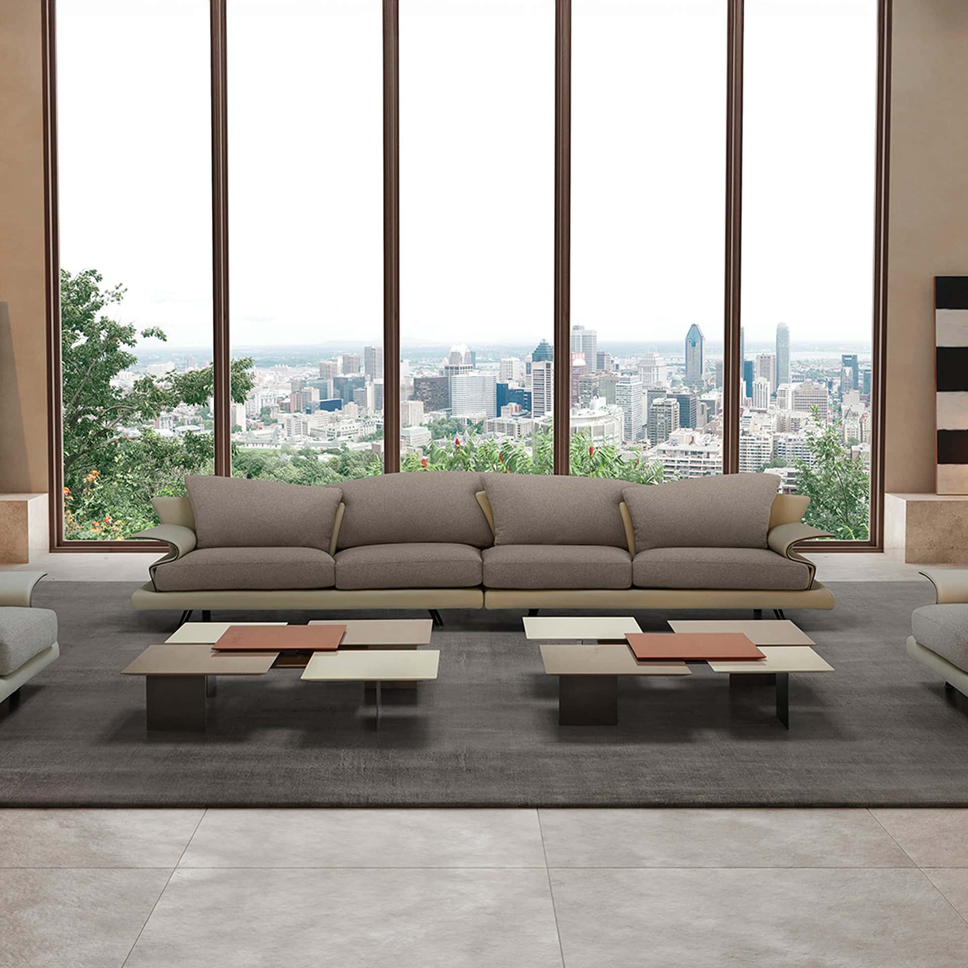 A modern living room with a long taupe sofa and two multi-level coffee tables, facing a large window that overlooks a sprawling city skyline with green trees in the foreground.