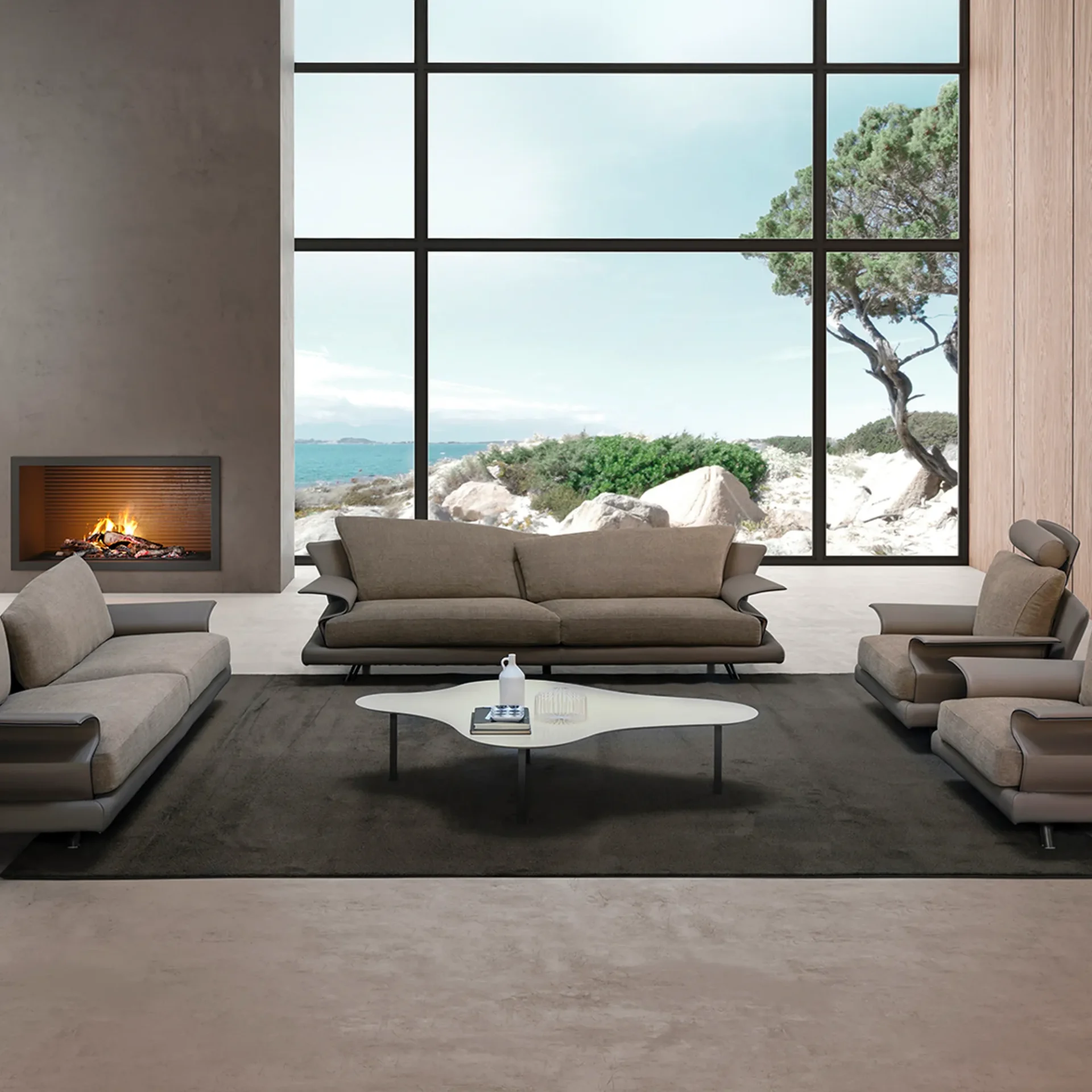 A modern living room with large windows overlooking a rocky coast and ocean. It features two light brown sofas, a white coffee table, and a fireplace.