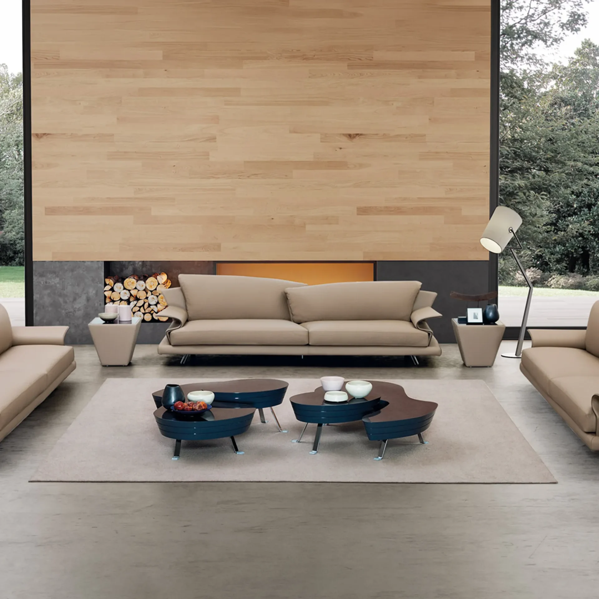 A modern living room with a beige sofa, unique dark blue and brown organic coffee tables on a light rug, and a fireplace with wood and an amber glow. A large window shows trees outside.