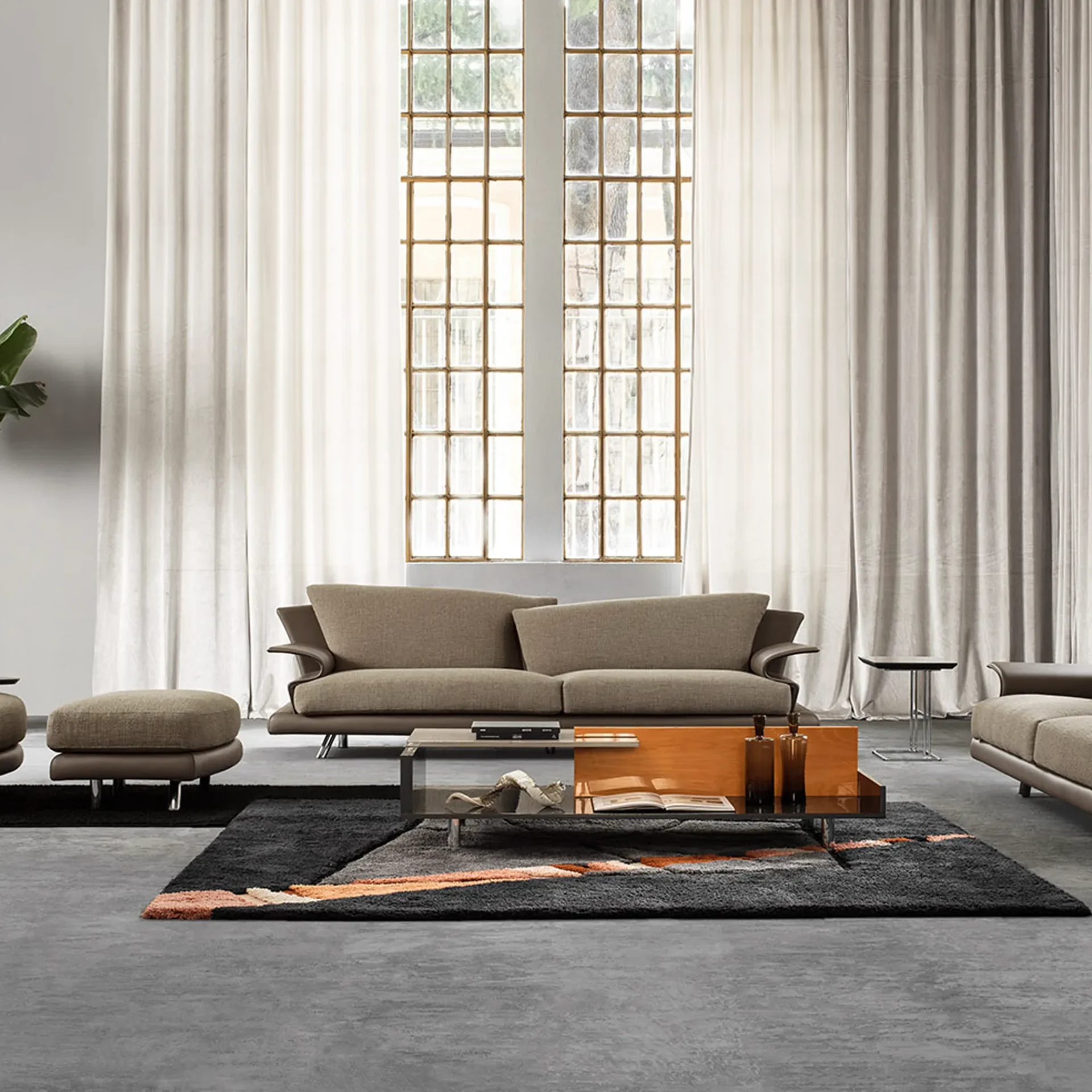 A modern living room featuring a beige sofa, matching ottoman, and a dark gray shaggy rug with orange abstract patterns. Large industrial-style windows with light cream curtains are in the background, and a multi-material coffee table sits in the foreground.