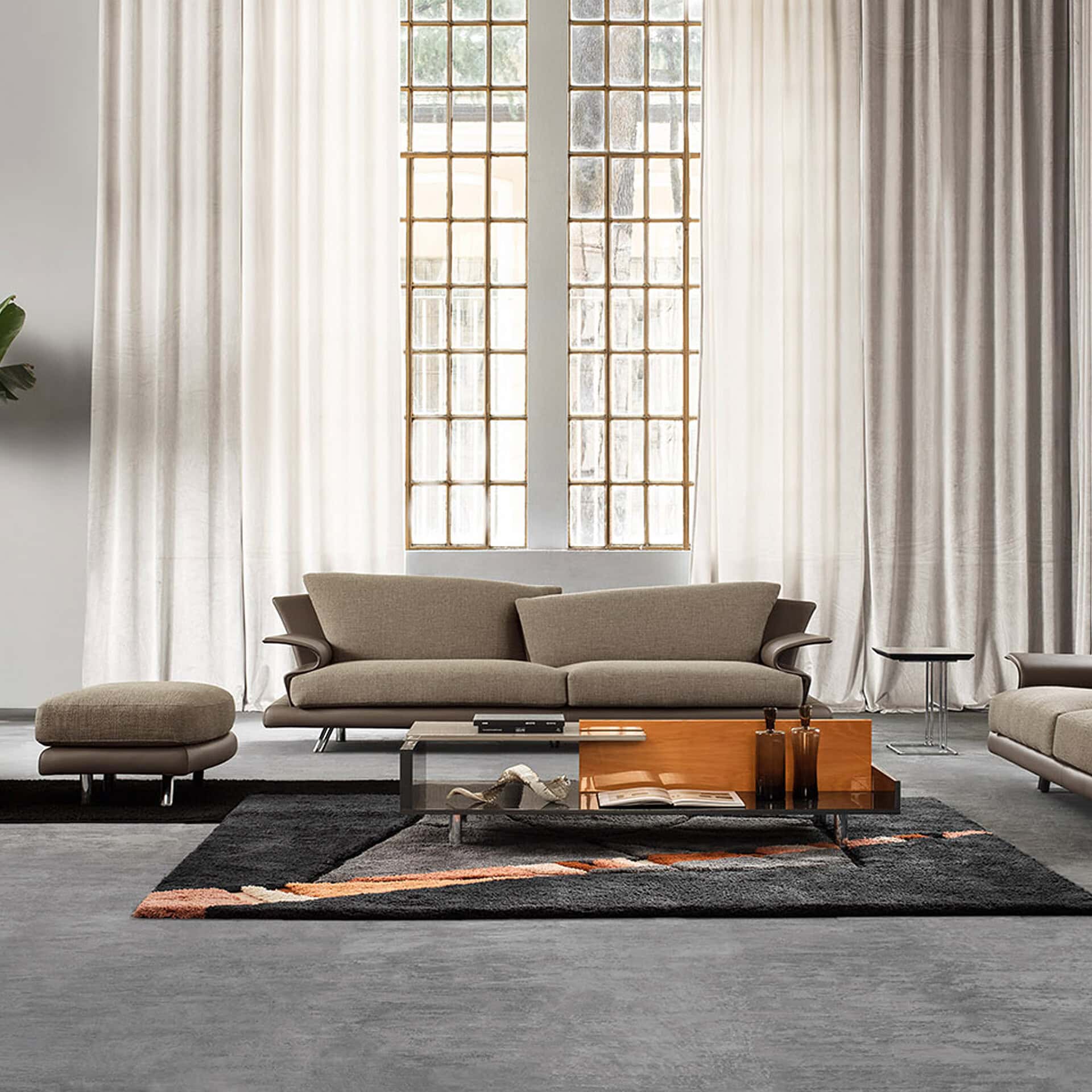 A modern living room featuring a beige sofa, matching ottoman, and a dark gray shaggy rug with orange abstract patterns. Large industrial-style windows with light cream curtains are in the background, and a multi-material coffee table sits in the foreground.