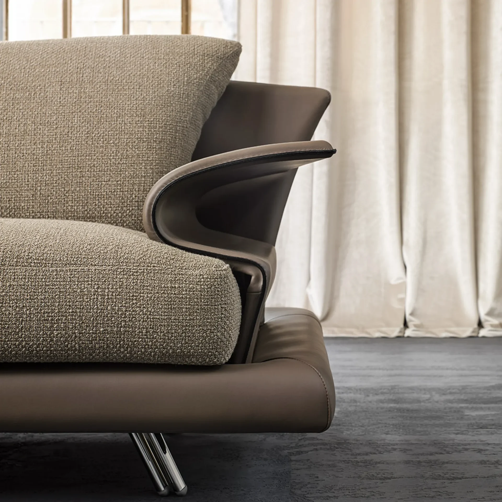 Close-up of a modern sofa with a textured light beige fabric cushion and a curved dark taupe leather armrest, set against a blurred background of cream curtains and a window.