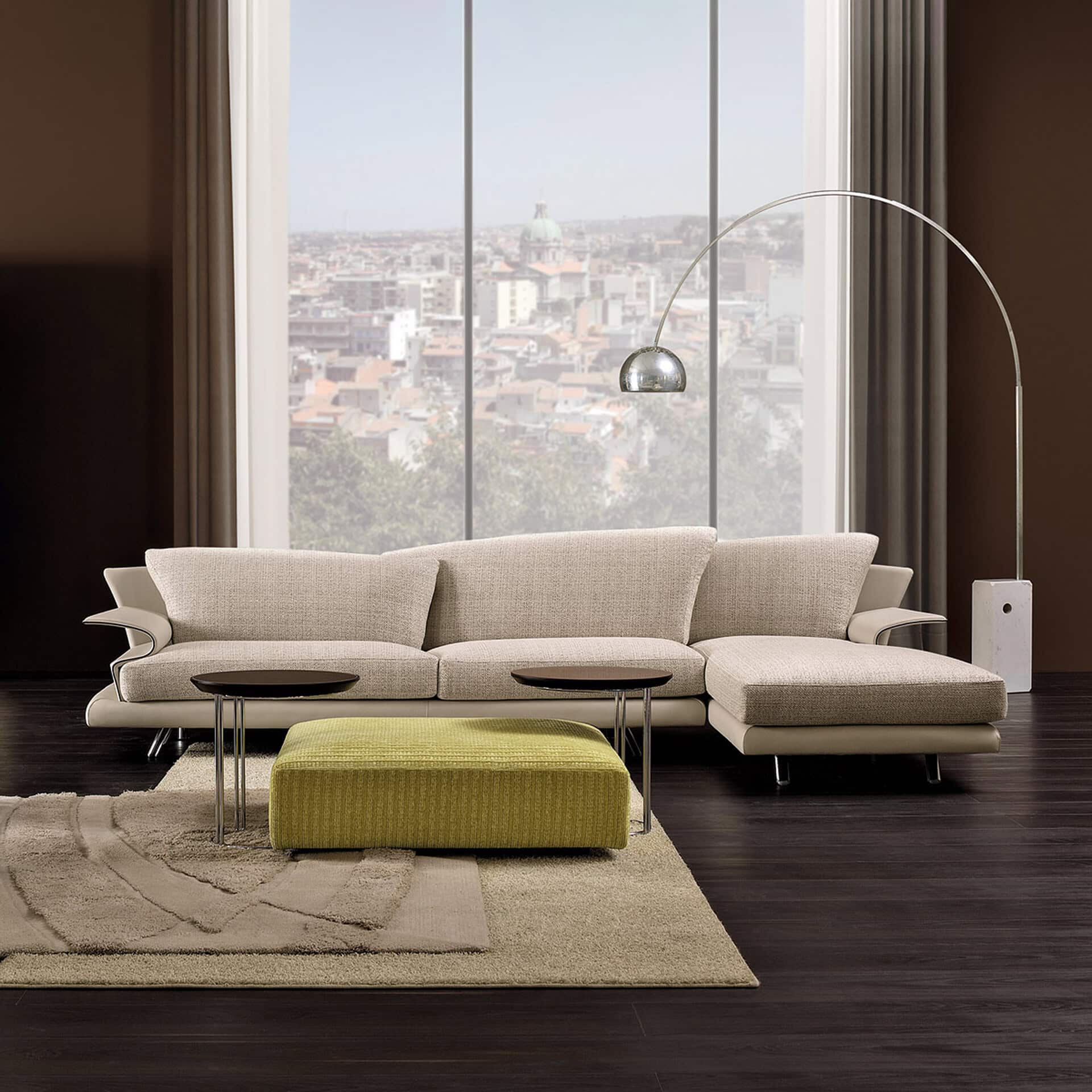 A modern living room with a beige sectional sofa, a lime green ottoman, and an arched floor lamp, set against a large window overlooking a city skyline.