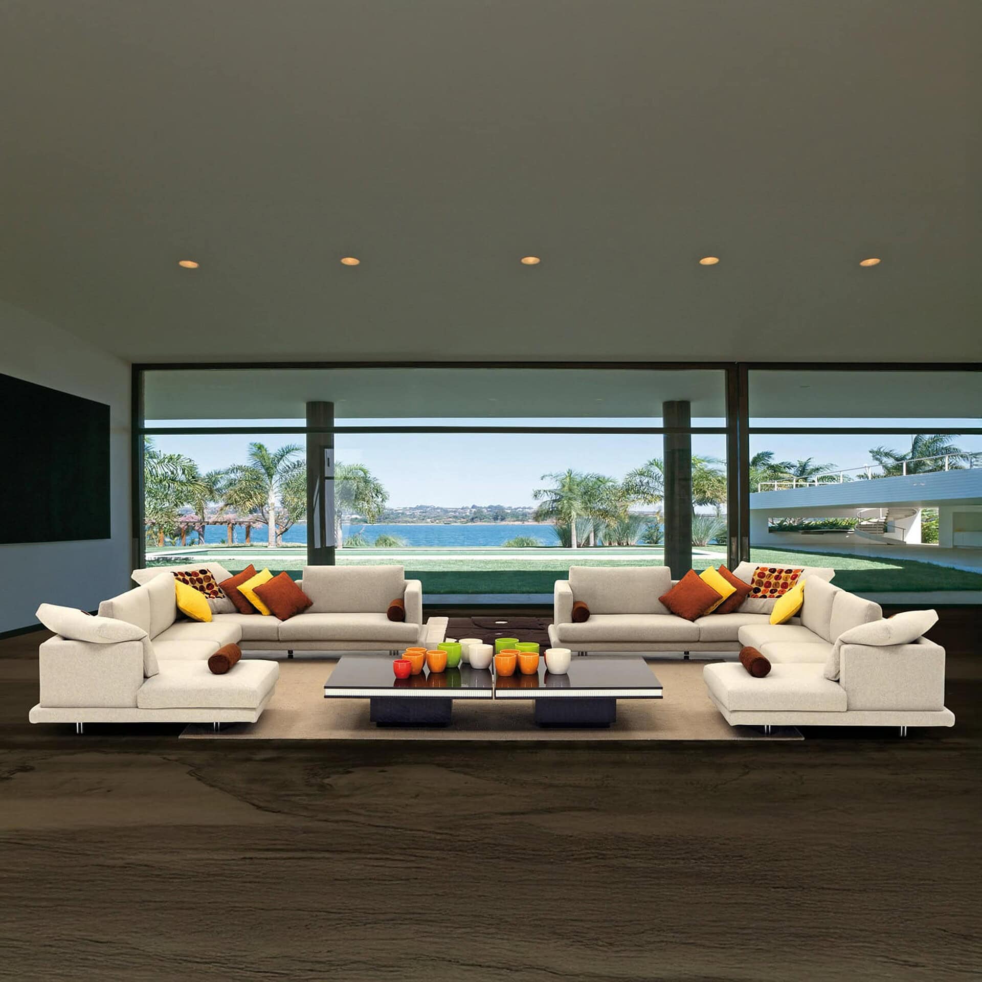 Luxurious modern living room with beige sofas, colorful pillows, and scenic water view through large glass windows.