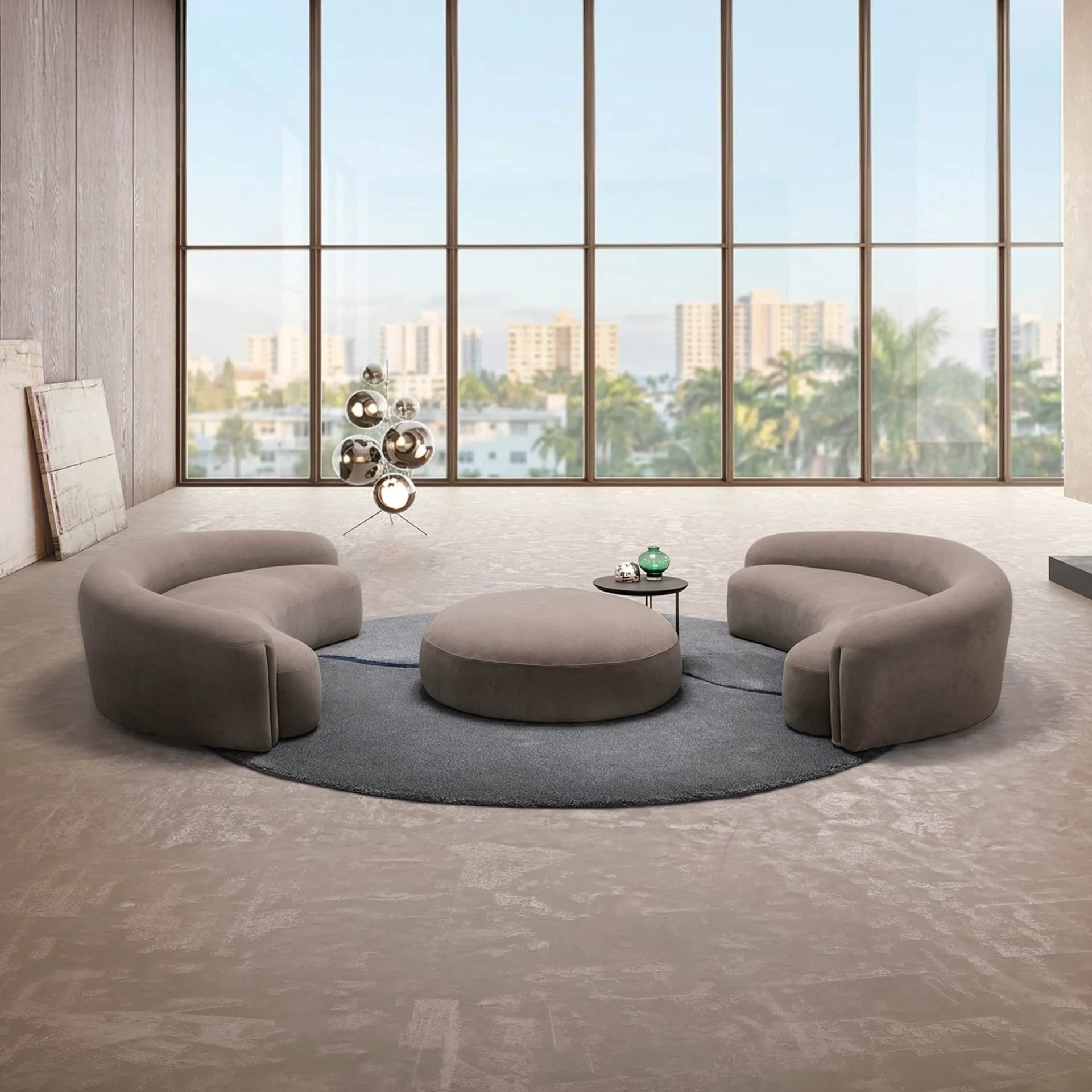 A modern living room with floor-to-ceiling windows showing a city view. Two curved taupe sofas and a round ottoman sit on a grey rug. A sculptural lamp and abstract art are also visible.