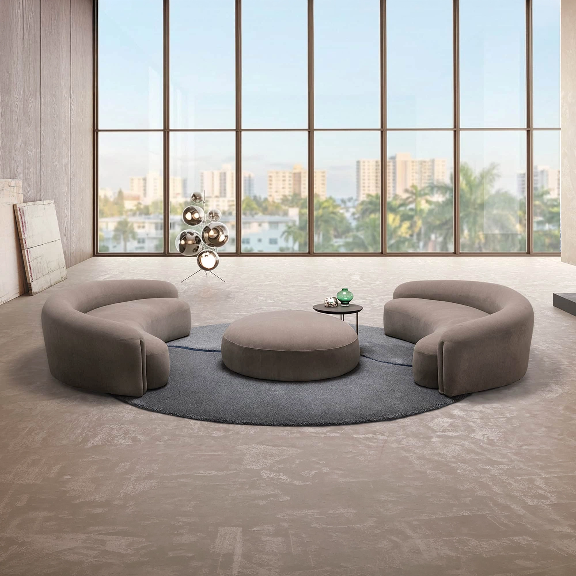 A modern living room with floor-to-ceiling windows showing a city view. Two curved taupe sofas and a round ottoman sit on a grey rug. A sculptural lamp and abstract art are also visible.
