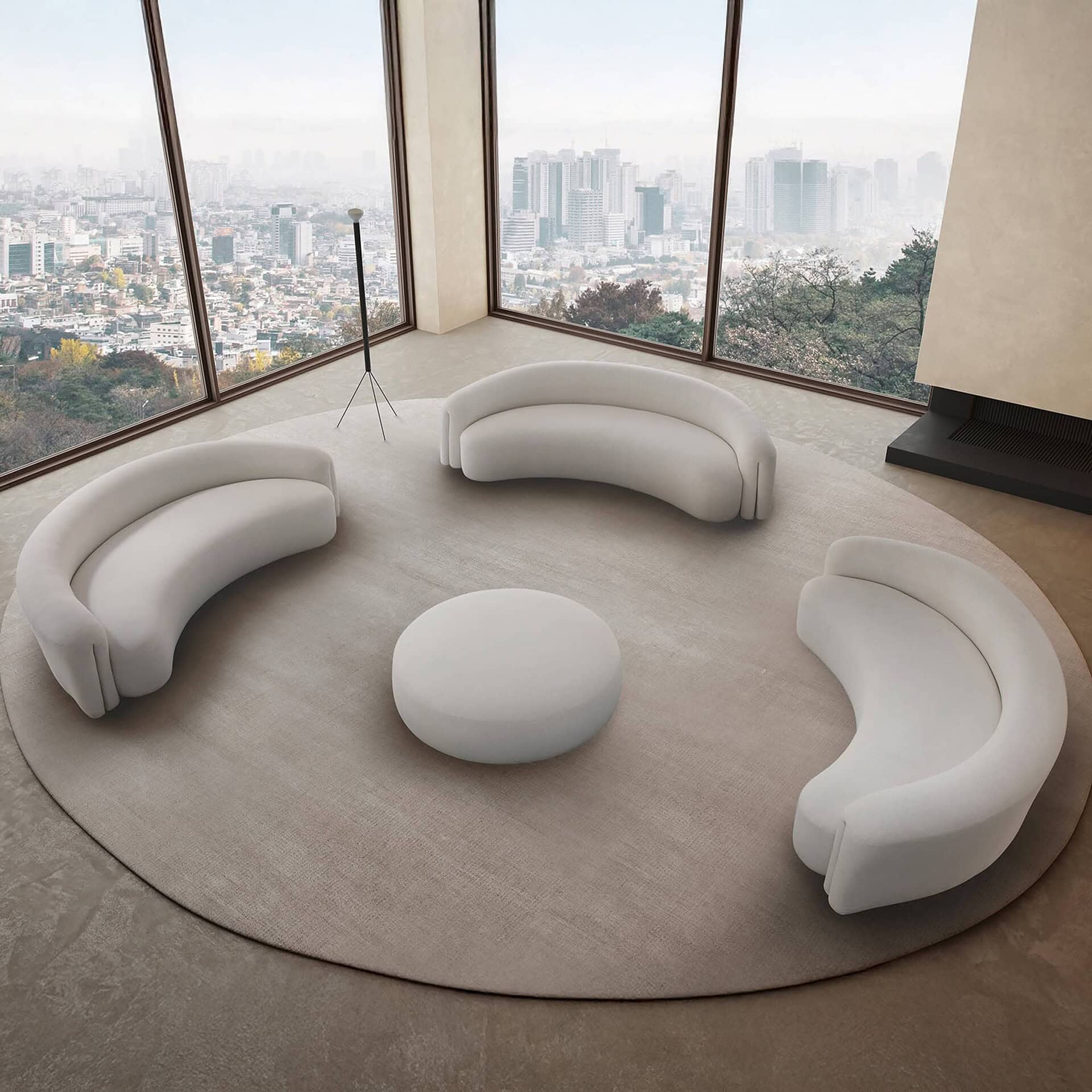 A modern living room with three curved white sofas and a round ottoman on a circular rug, facing floor-to-ceiling windows with a panoramic view of a hazy city skyline.