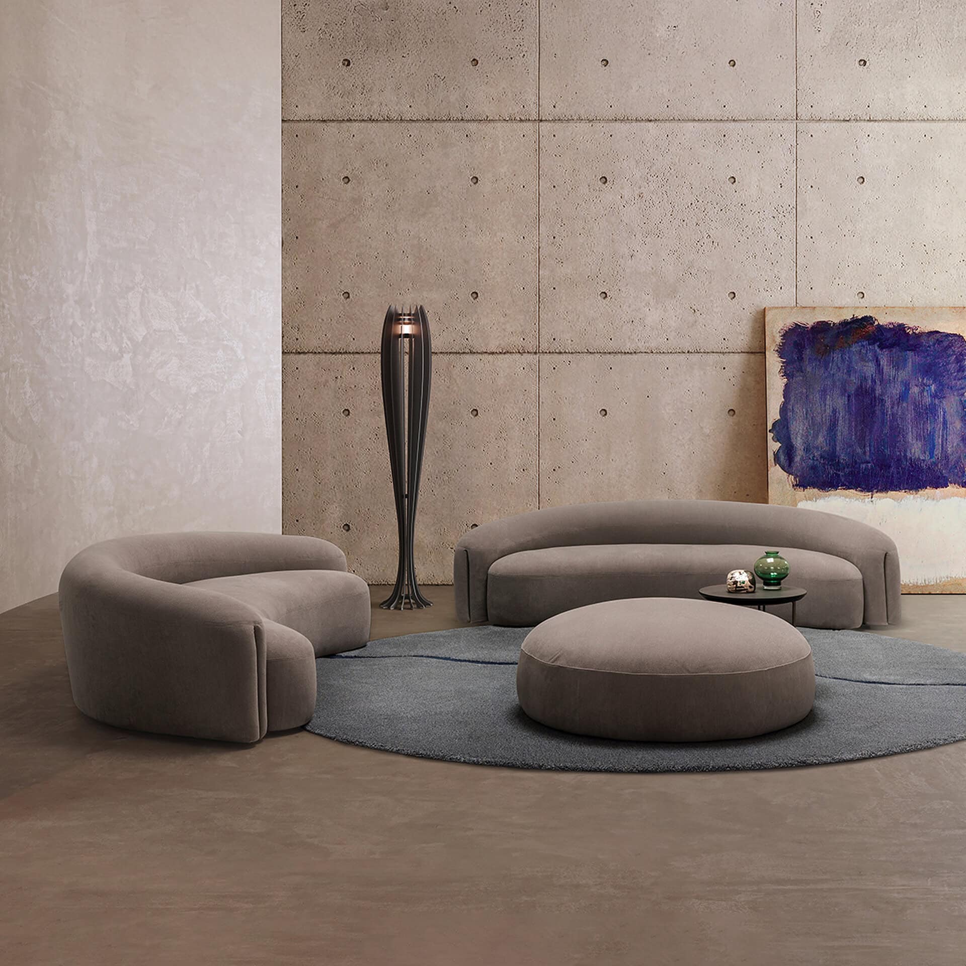 A modern living room with two curved taupe sofas, a matching ottoman, and a round gray rug on a concrete floor. A tall, dark sculptural lamp stands against an exposed concrete wall, next to which an abstract blue and white painting leans. The opposite wall is light textured plaster.
