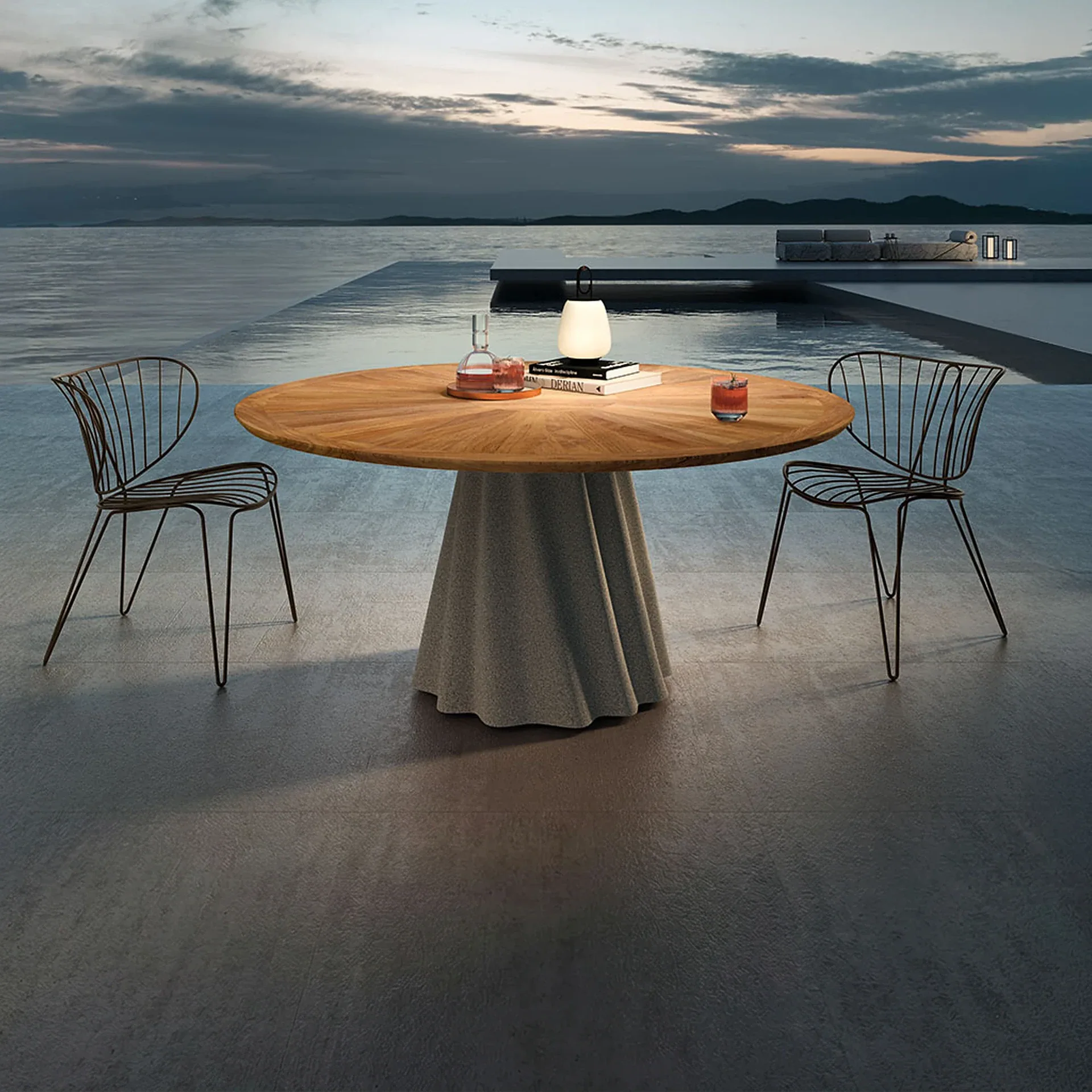 An outdoor dining scene at dusk with a round wooden table, two wire chairs, a glowing lamp, and drinks on a patio overlooking a body of water with distant land.