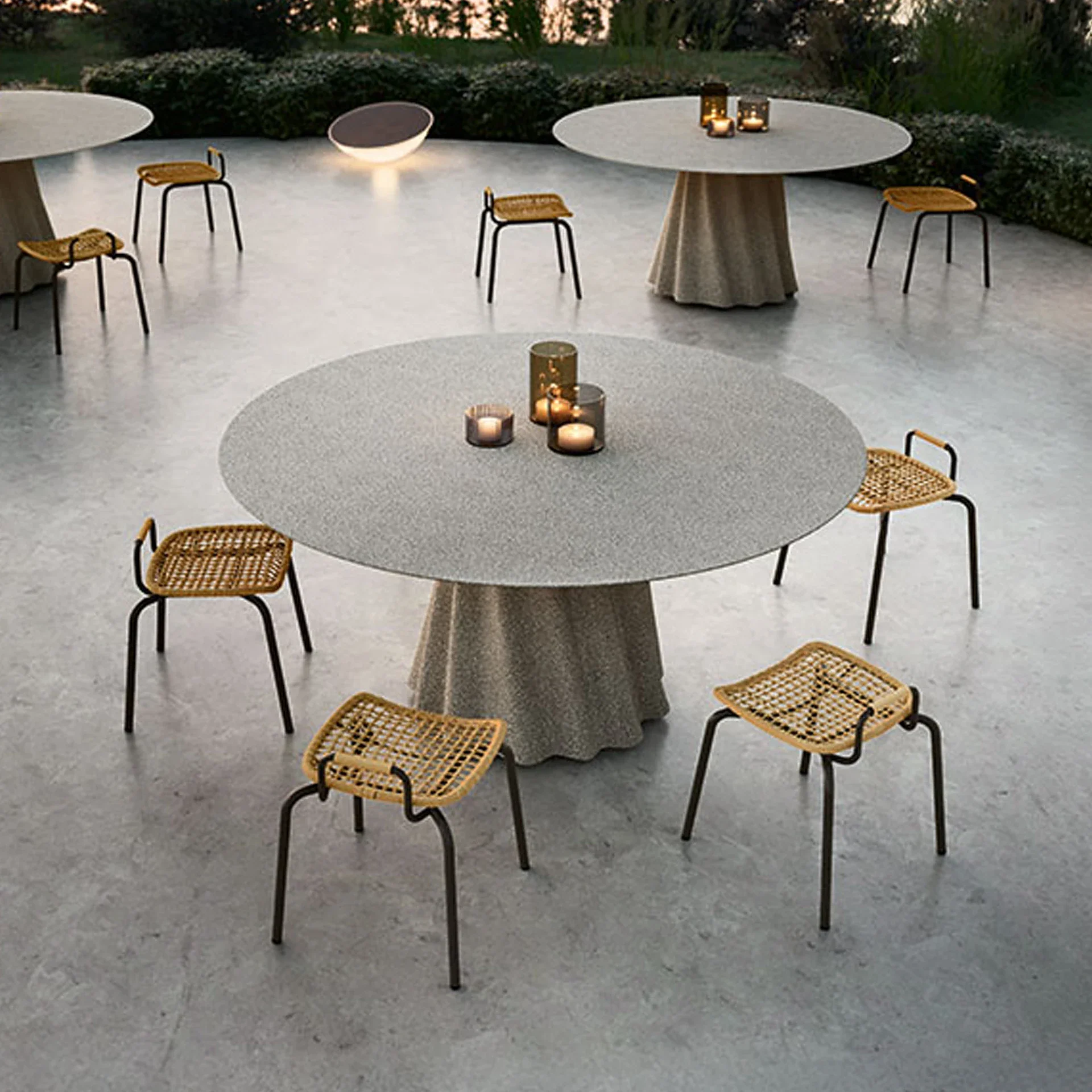 An outdoor patio with three round tables, woven rattan stools, and lit candles, set against a backdrop of dark green foliage at dusk.
