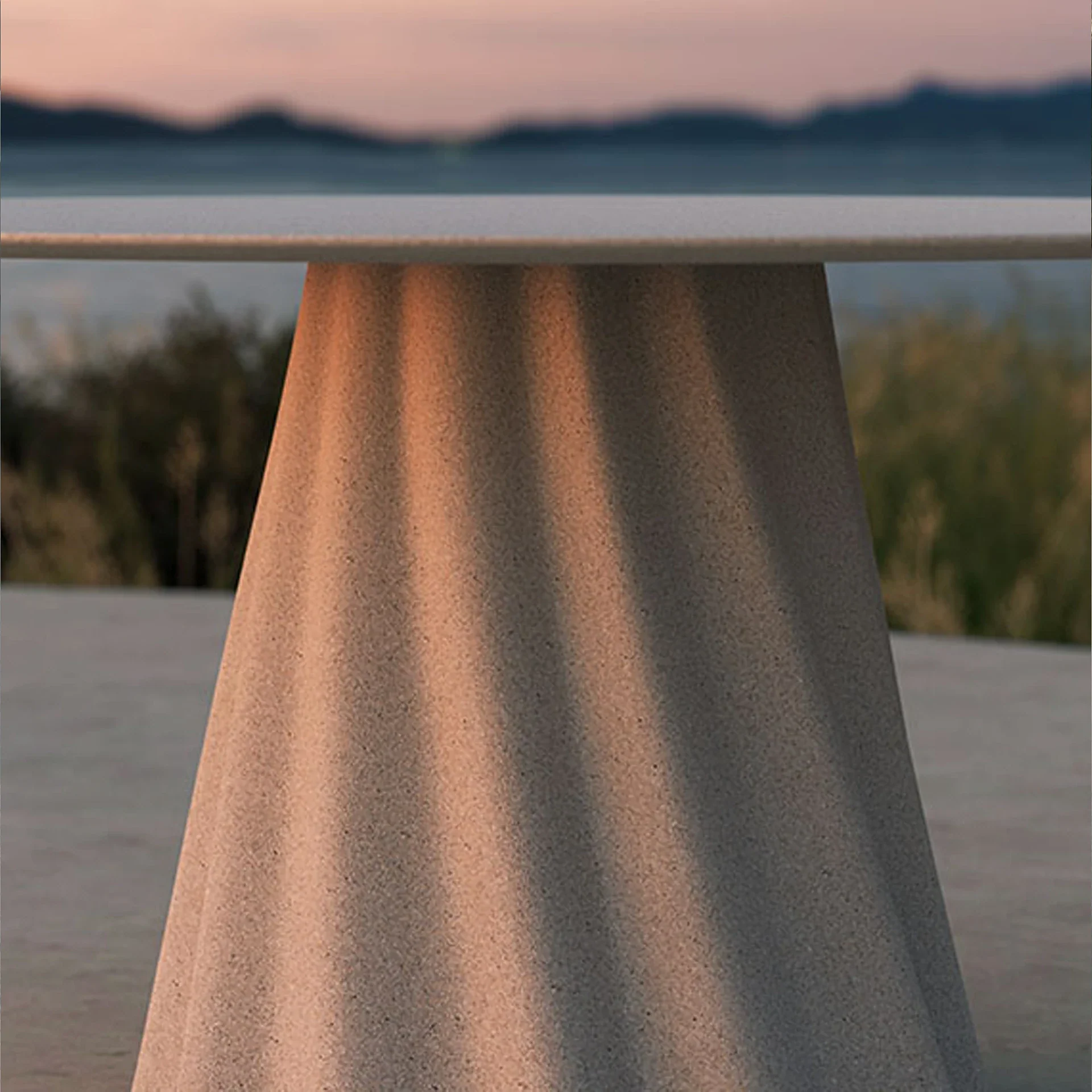A modern outdoor table with a textured, conical base and a flat, light-colored top, set against a blurred sunset landscape with mountains and water.