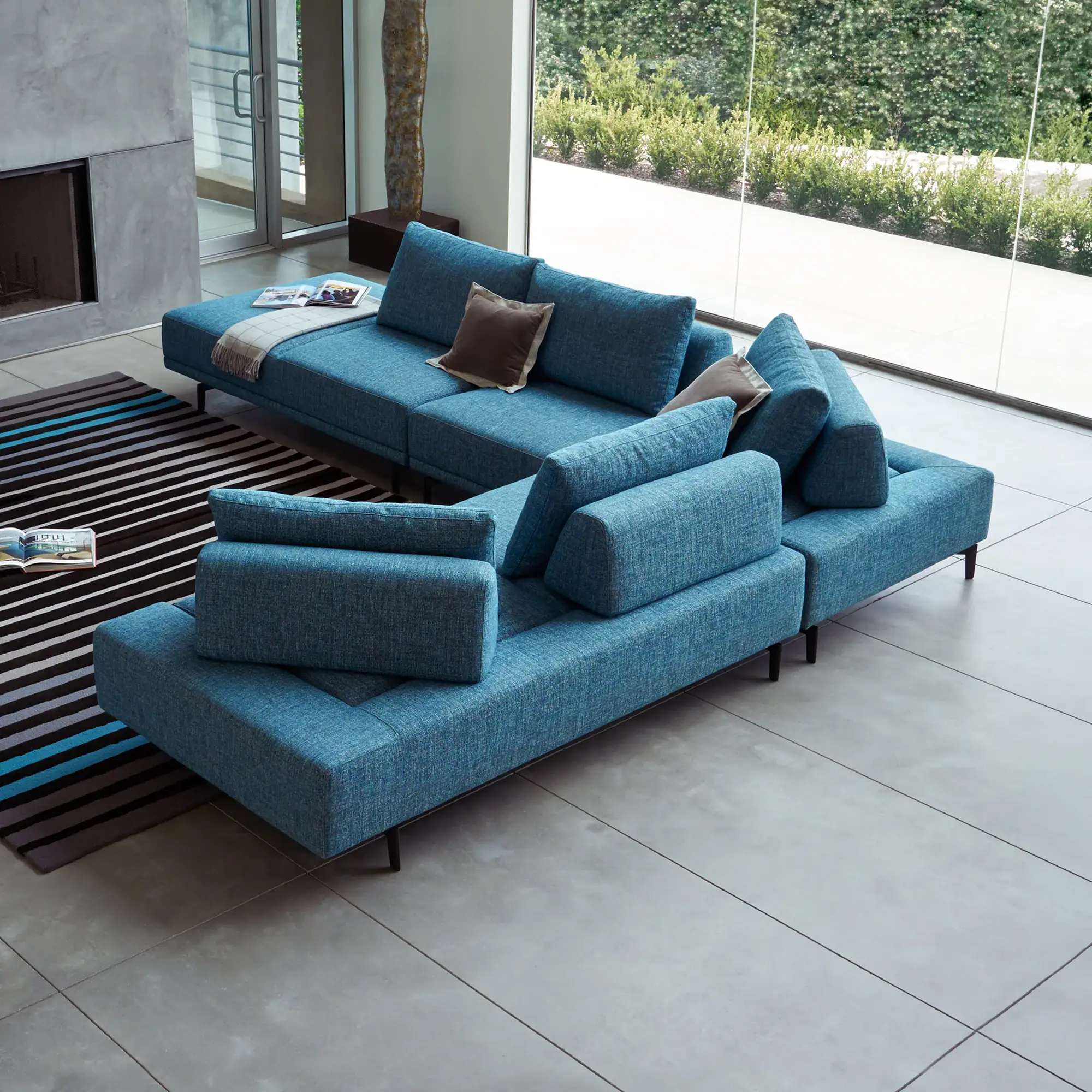 A modern living room with a large blue sectional sofa, a gray fireplace, and floor-to-ceiling windows revealing a green garden.