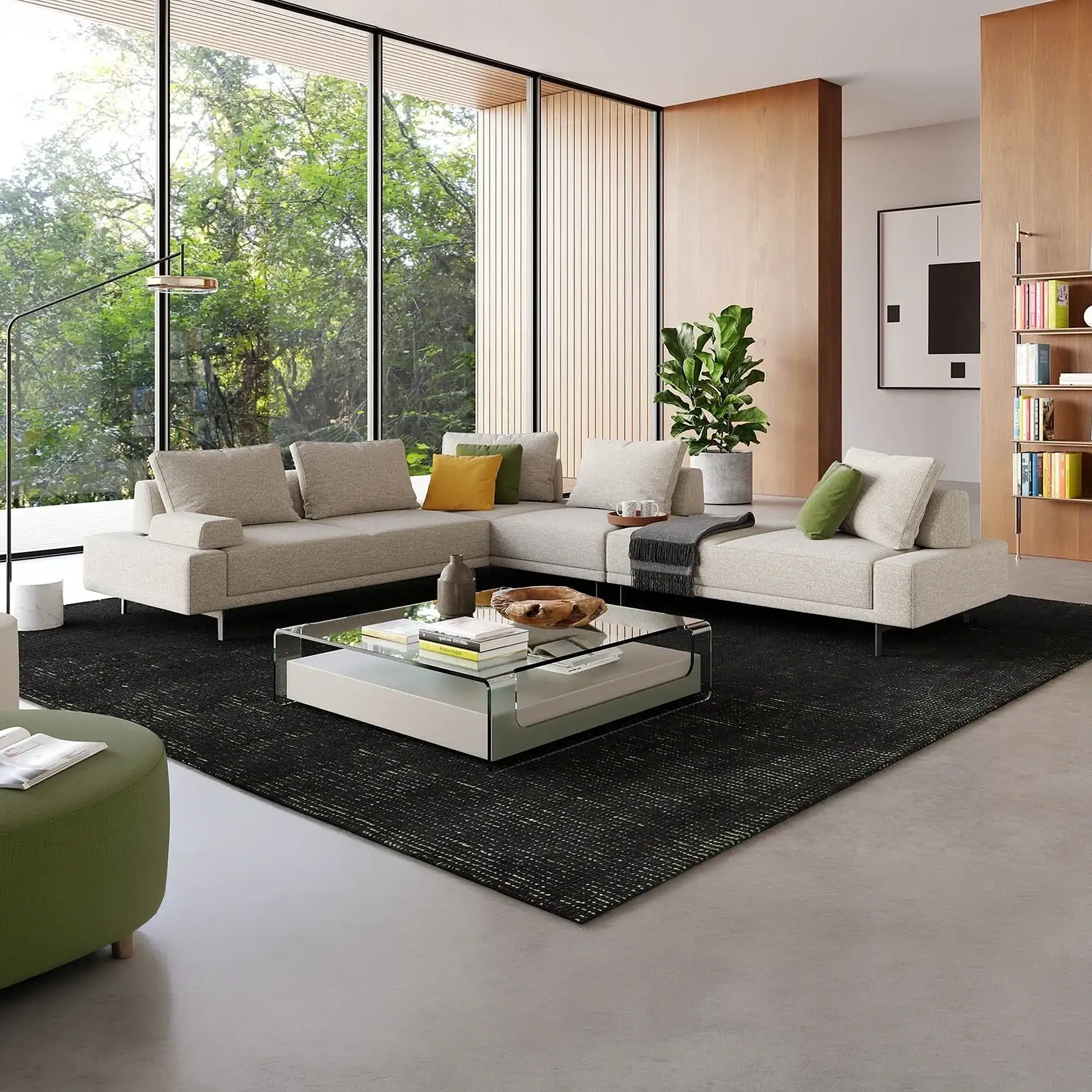 A modern living room with a light beige L-shaped sofa, glass coffee table, dark rug, and floor-to-ceiling windows overlooking green trees.