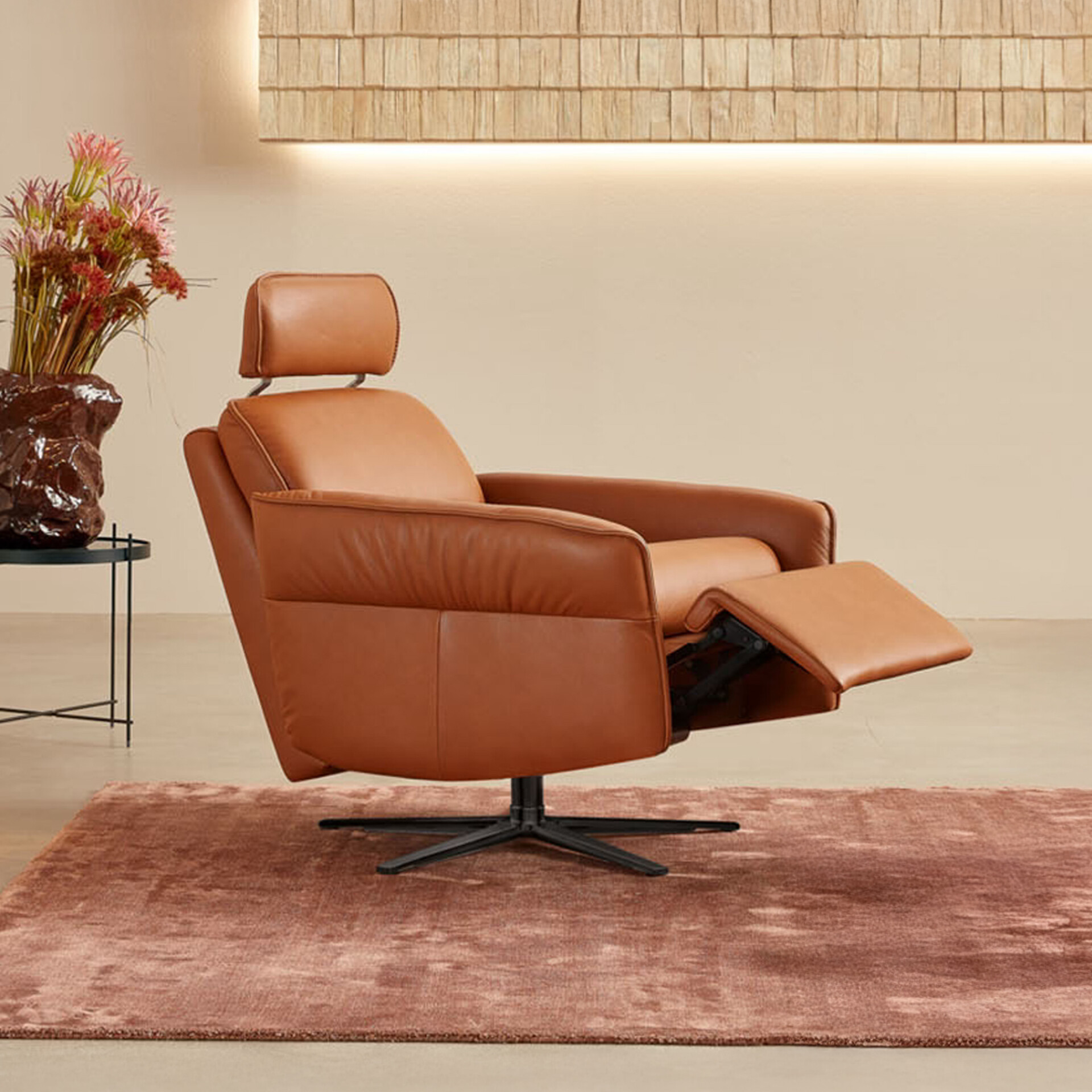 A modern caramel leather recliner with an extended footrest and adjustable headrest, sitting on a black swivel base on a dusty rose rug. A wooden shingle wall panel with warm lighting is behind it, and a side table with a floral arrangement is to the left.