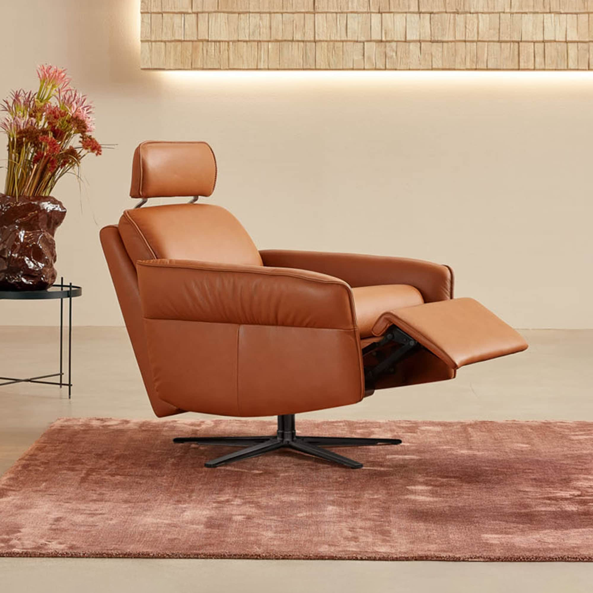 A modern caramel leather recliner with an extended footrest and adjustable headrest, sitting on a black swivel base on a dusty rose rug. A wooden shingle wall panel with warm lighting is behind it, and a side table with a floral arrangement is to the left.