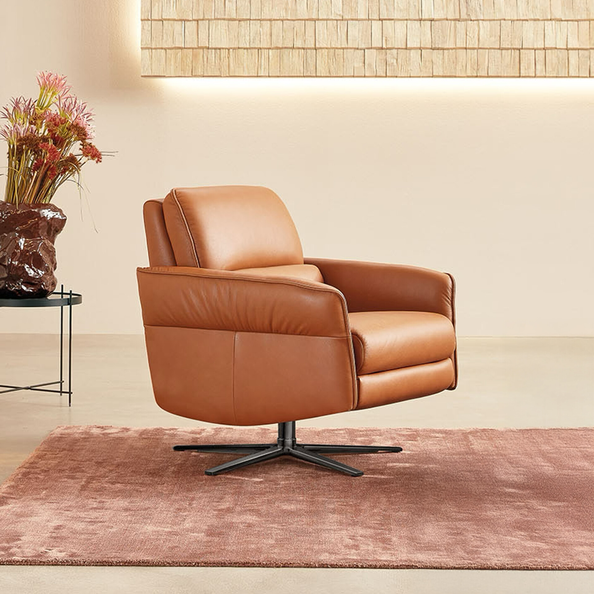 A tan leather swivel armchair with a black base sits on a dusty rose rug in a modern room. A side table with a vase of dried flowers is to the left, and a textured wall panel with integrated lighting is in the background.