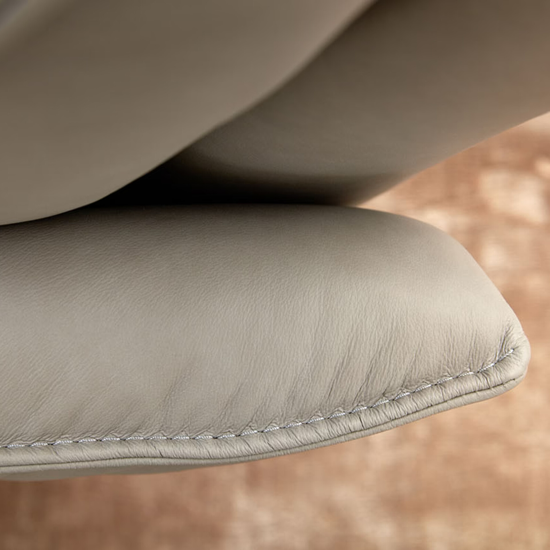 Close-up of a light gray leather furniture cushion with visible stitching and subtle texture, against a blurred warm background.