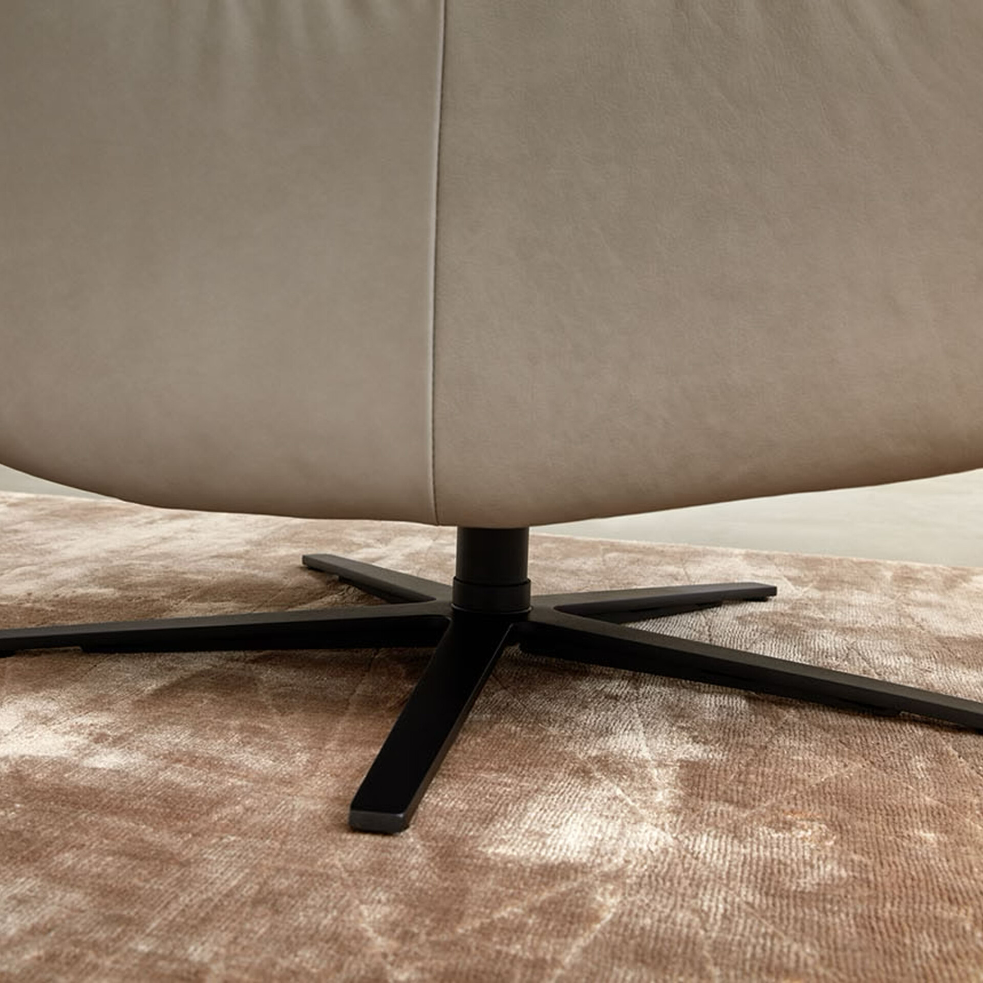 A close-up of a modern light beige armchair with a matte black five-star swivel base, sitting on a textured light brown rug.