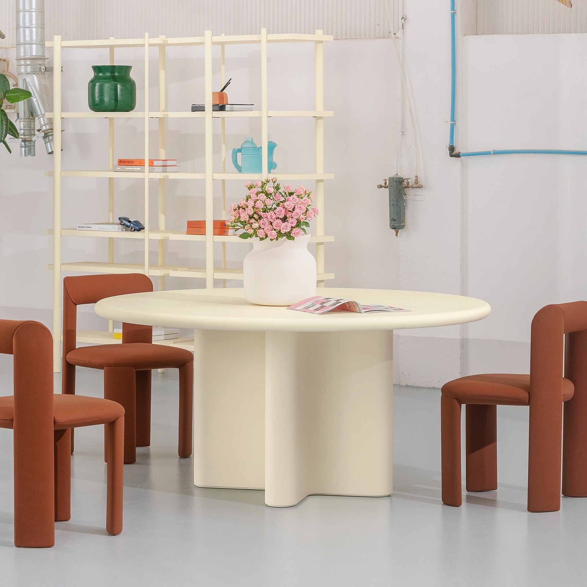 Modern round cream-colored dining table with four rust-colored upholstered chairs, decorated with a pink rose bouquet in a white vase.
