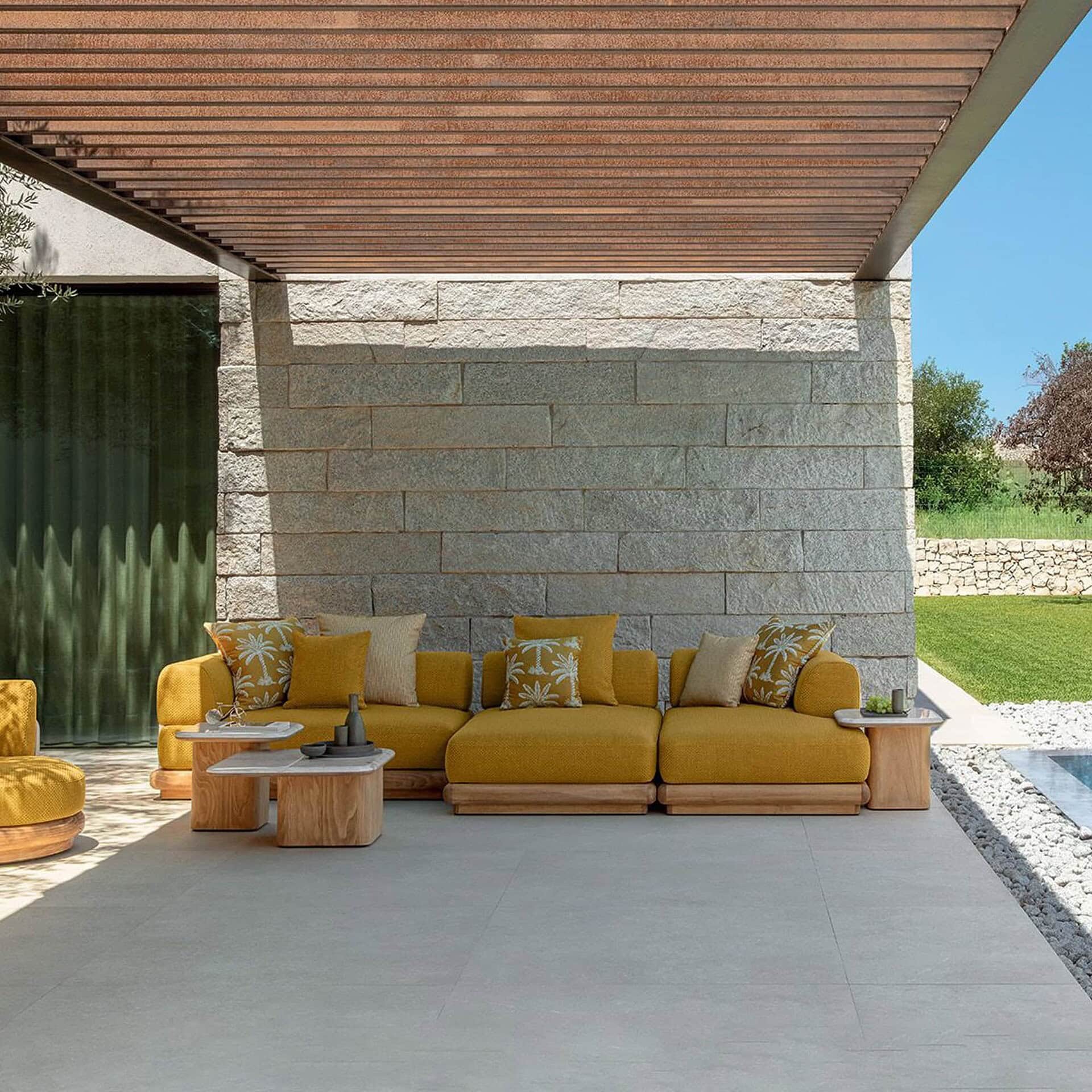 Modern outdoor living space with yellow sectional sofa, wooden coffee tables, and stone wall.