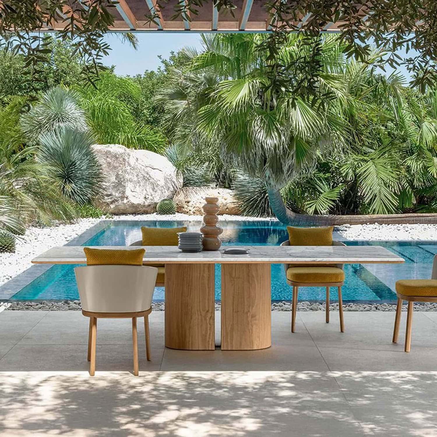 Elegant outdoor dining table with green cushions and natural wood chairs beside a pool, surrounded by lush tropical plants.