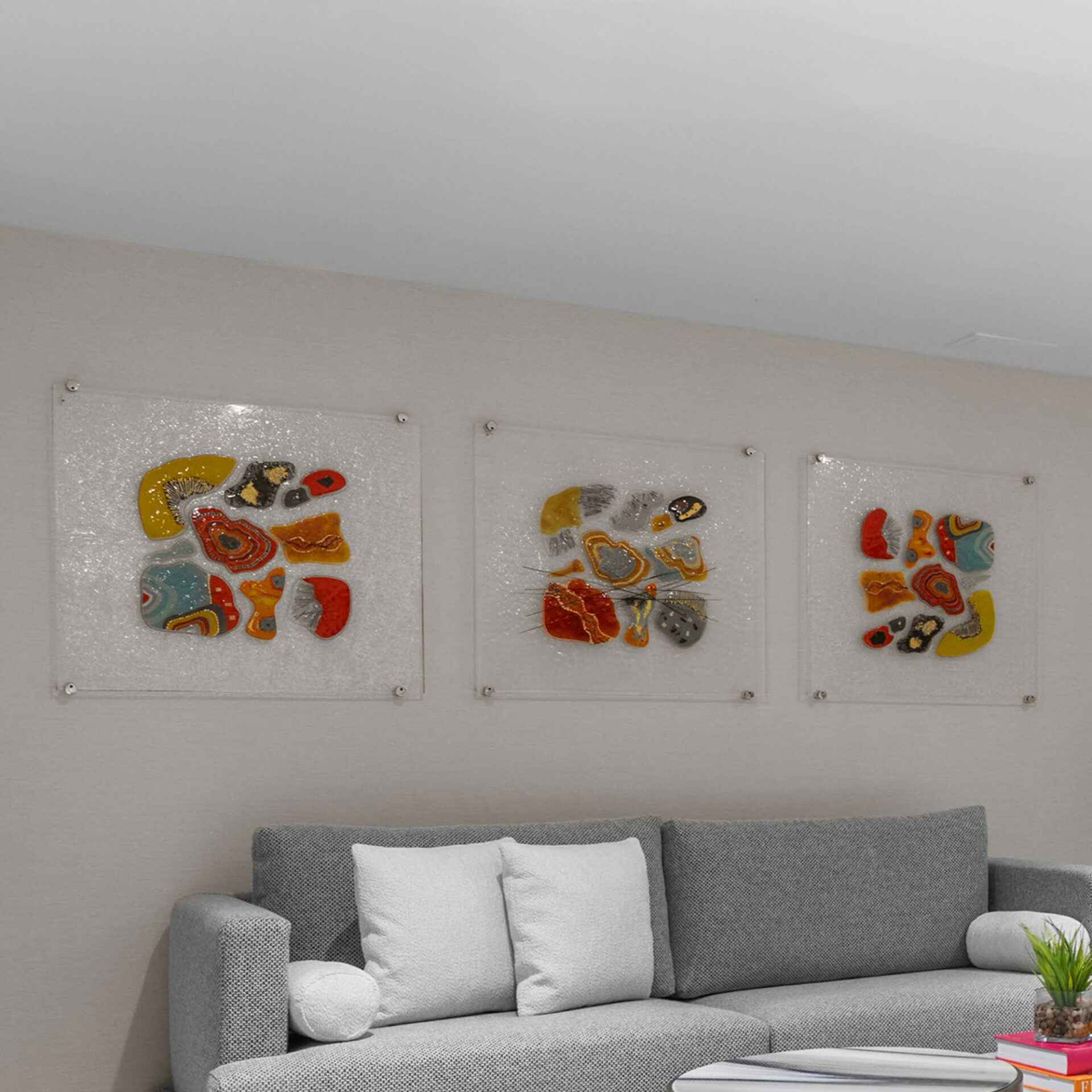 Three colorful abstract art pieces with organic shapes on clear panels mounted on a light gray wall above a modern gray sectional sofa with white pillows.