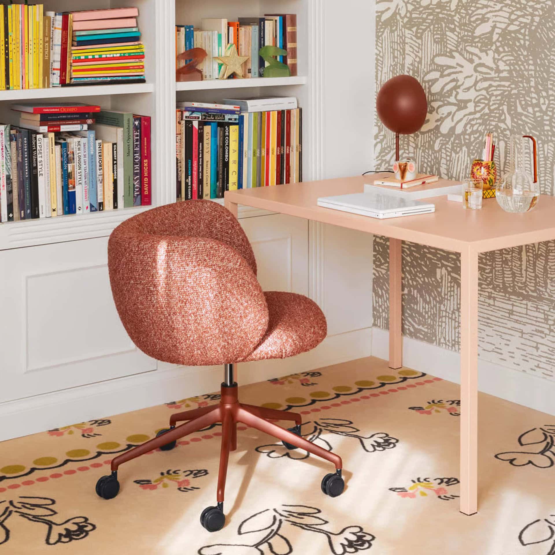 Modern pink desk and cozy upholstered office chair in a home study with colorful bookshelf and decorative accents.