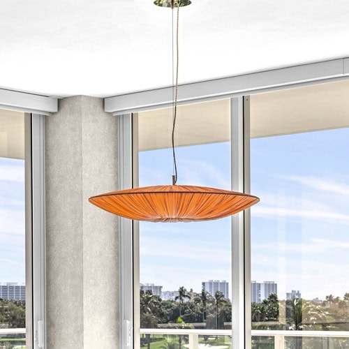 A modern, oval-shaped pendant light fixture with a cream-colored pleated fabric shade, emitting a warm, golden glow. It is suspended from a light ceiling by thin wires.