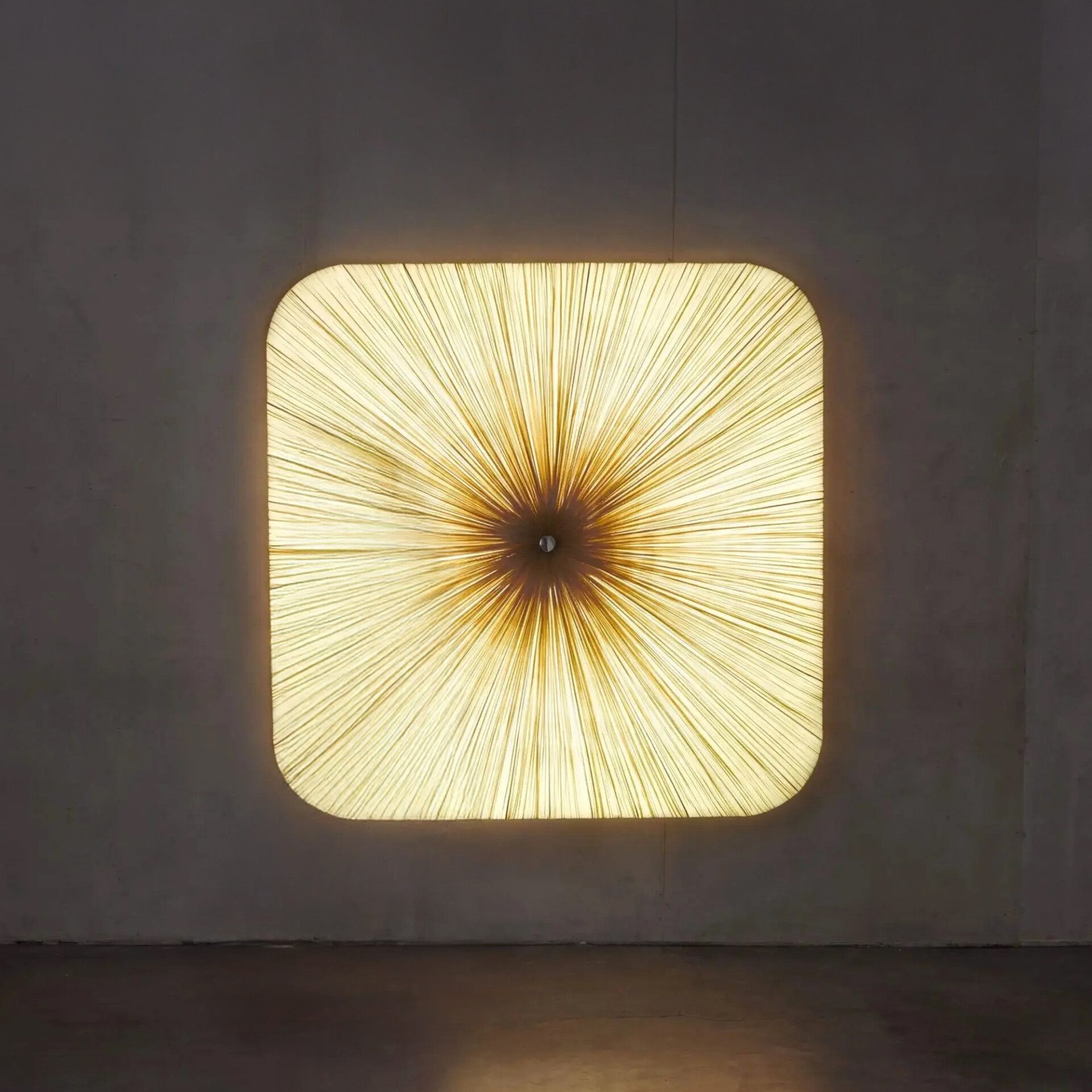 A square light fixture with rounded corners on a dark wall, emitting a warm, golden starburst pattern from its center.