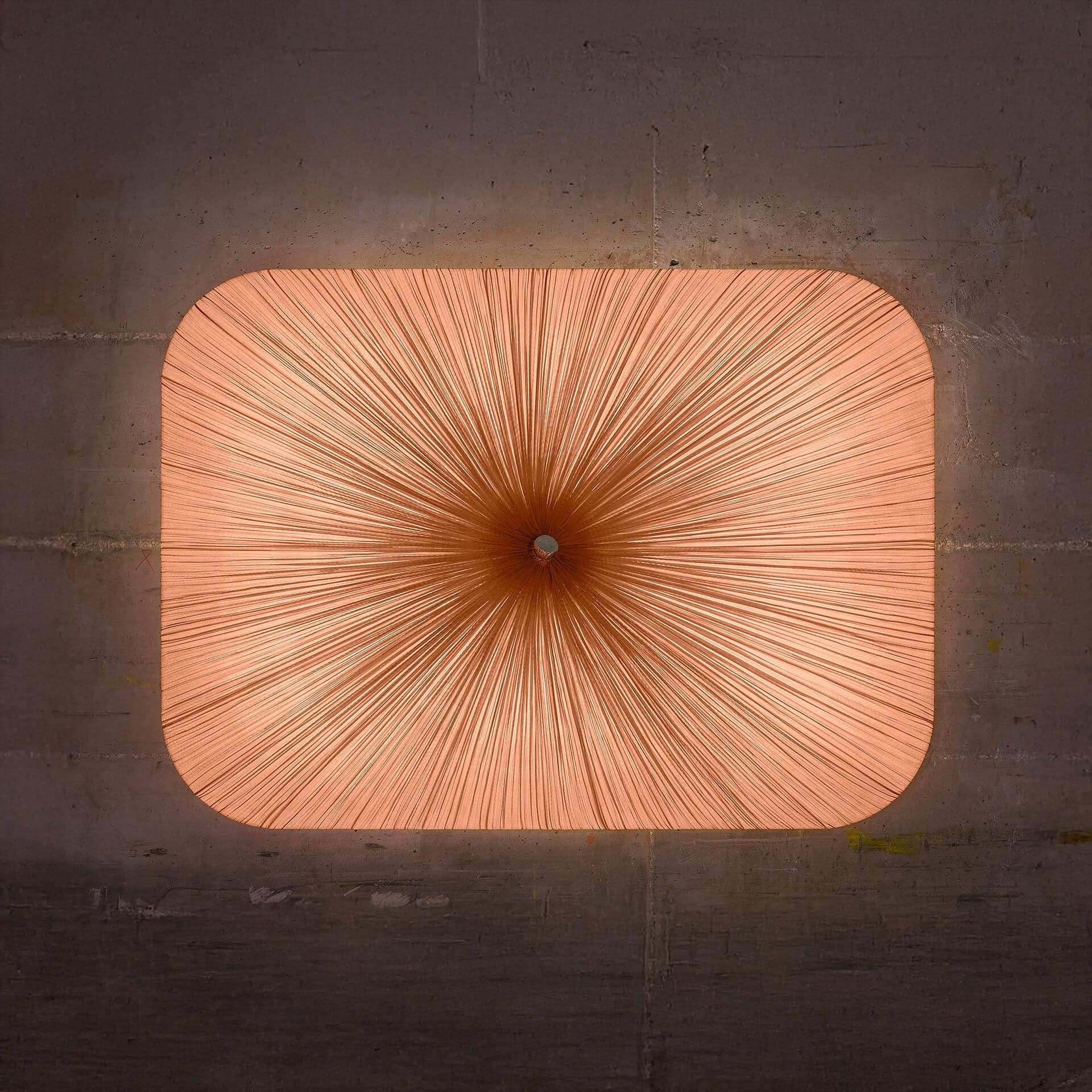 A square, rounded-corner light fixture with a radiating starburst pattern of thin, orange threads, set against a dark, textured concrete ceiling or wall.