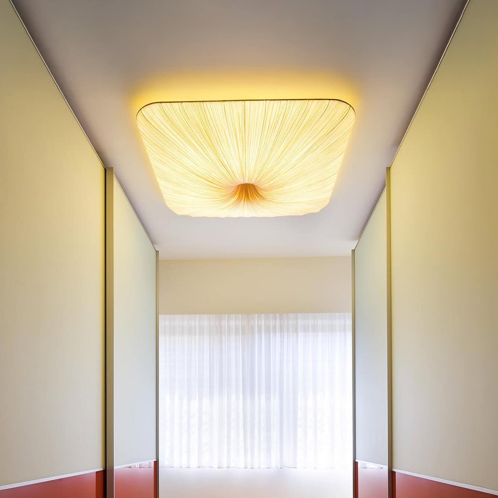 A modern hallway with a unique square pleated ceiling light emitting warm yellow light, leading to a window with sheer white curtains.