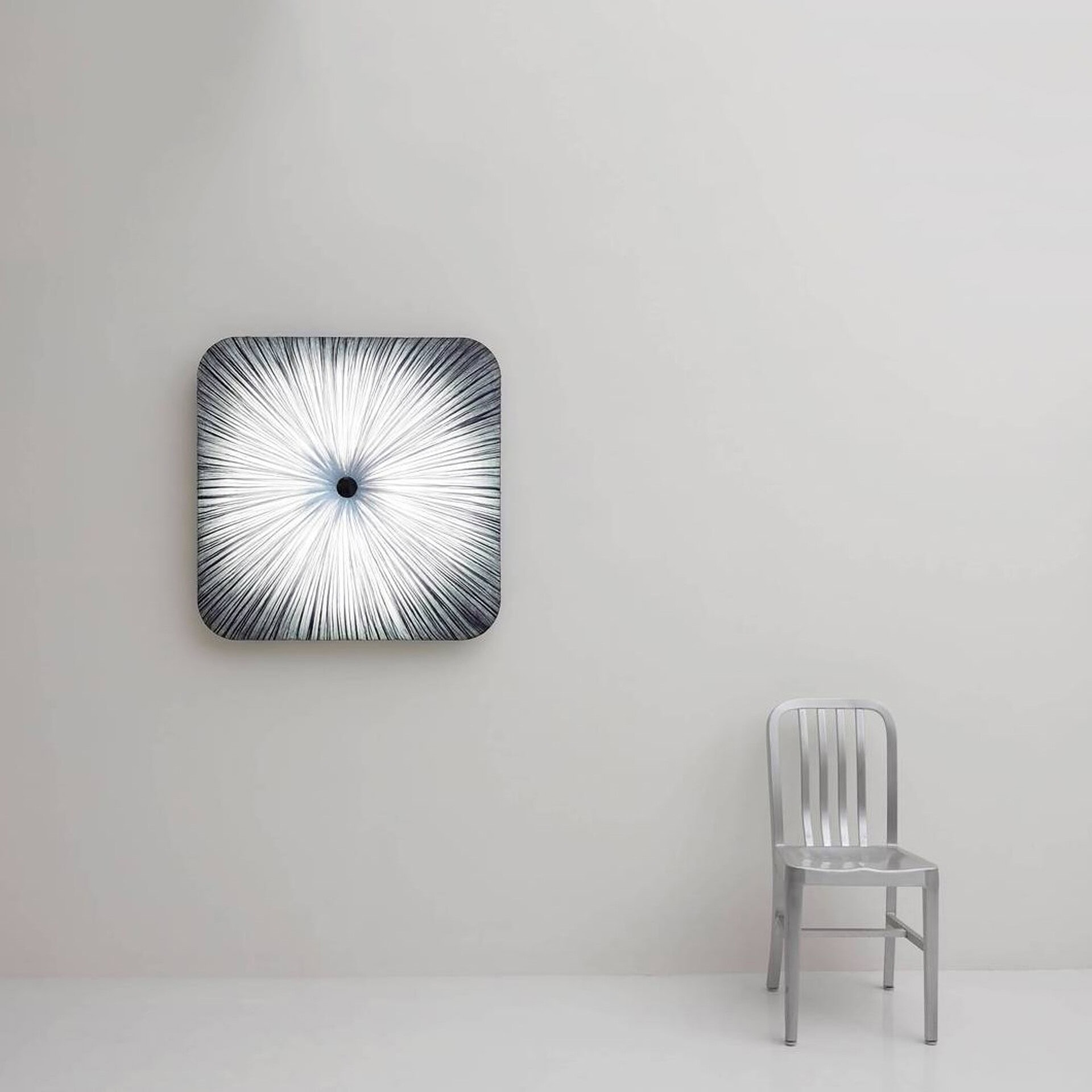 A square wall light with radiating white lines and a central dark circle, next to a silver metal chair, in a minimalist room.