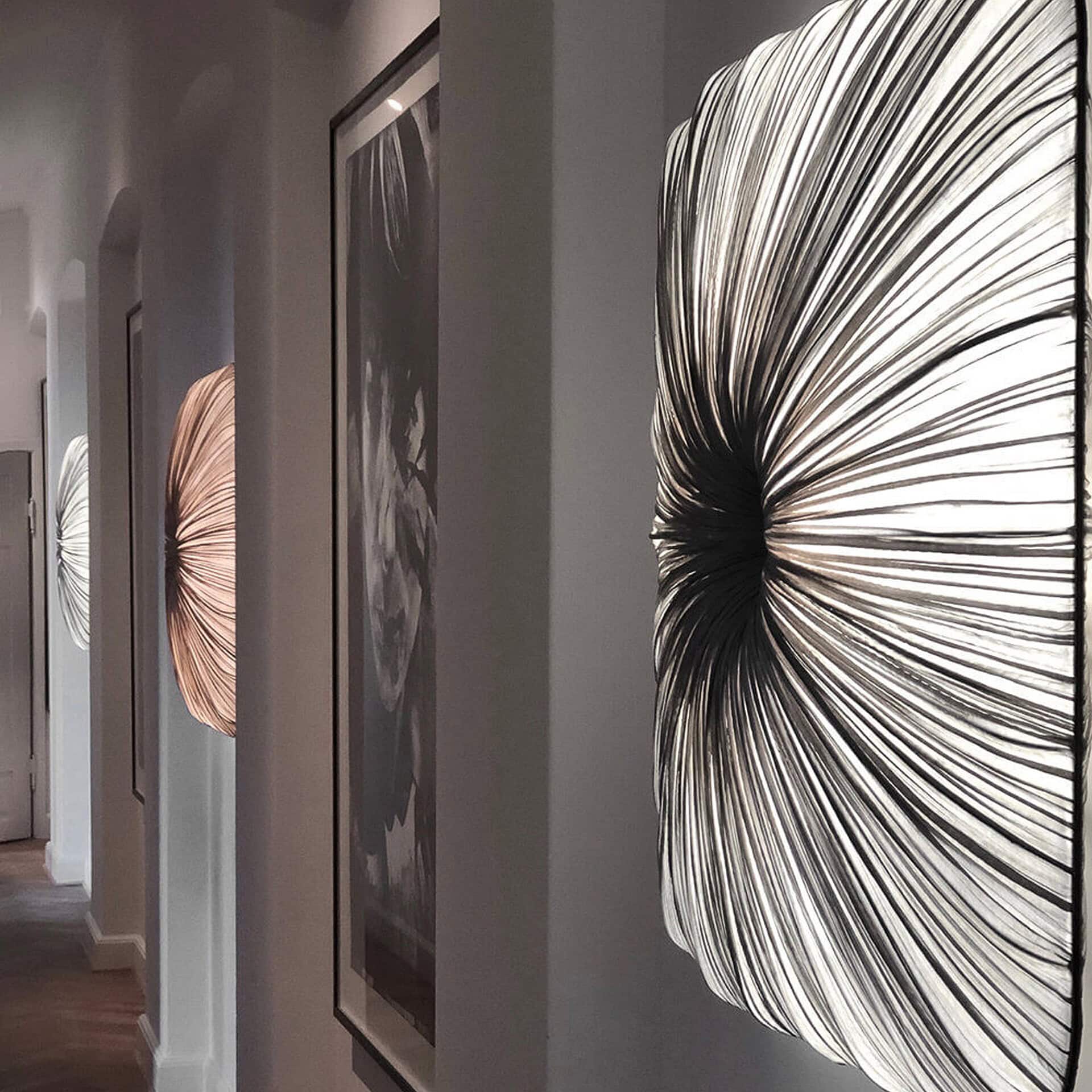 A modern hallway with three circular, pleated wall sconces glowing. The largest, in the foreground, is silver-white, while a smaller one further back is copper-toned. Framed art hangs on the walls.