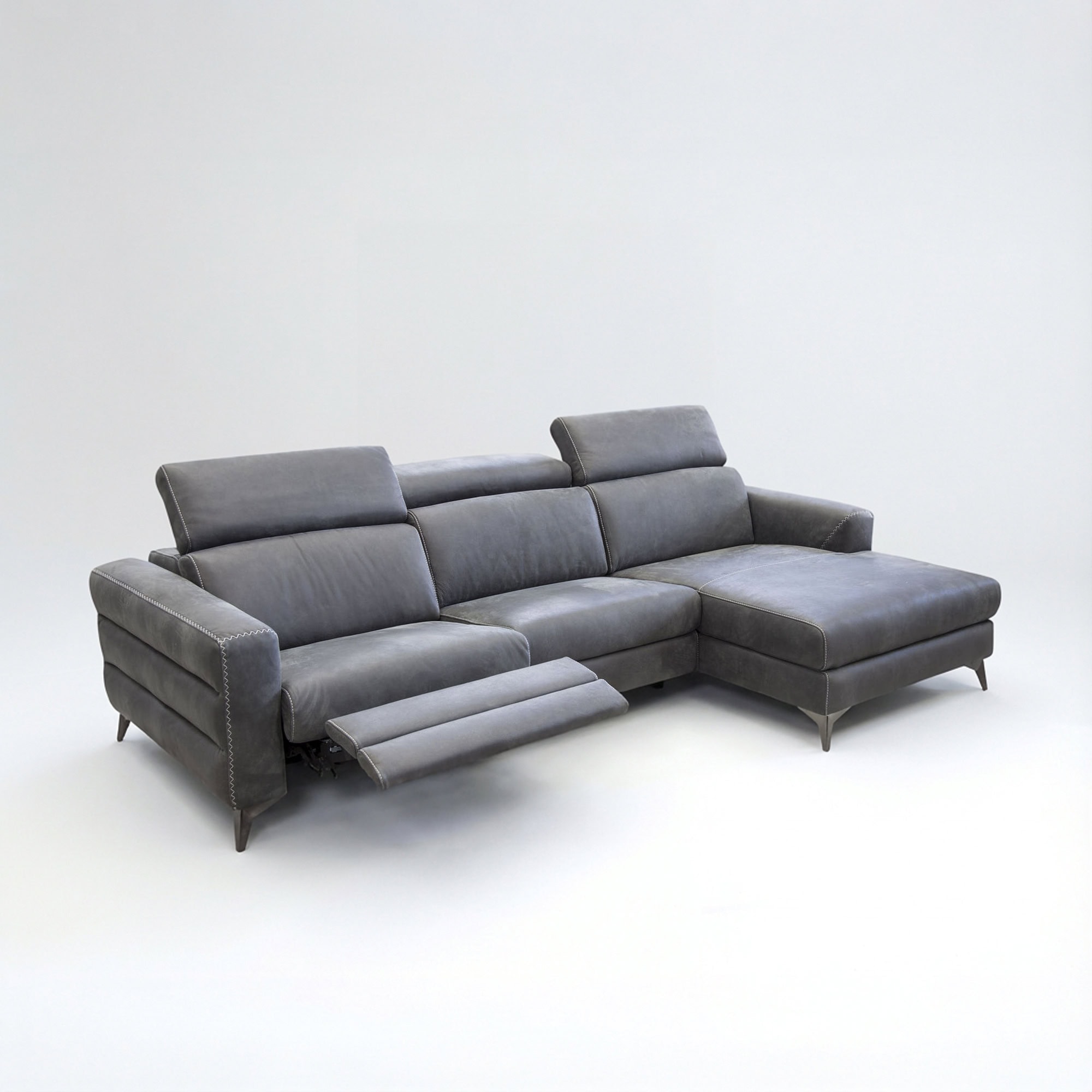 Dark grey L-shaped sectional sofa with an extended recliner footrest, adjustable headrests, and a chaise lounge, featuring white contrast stitching, on a light grey background.