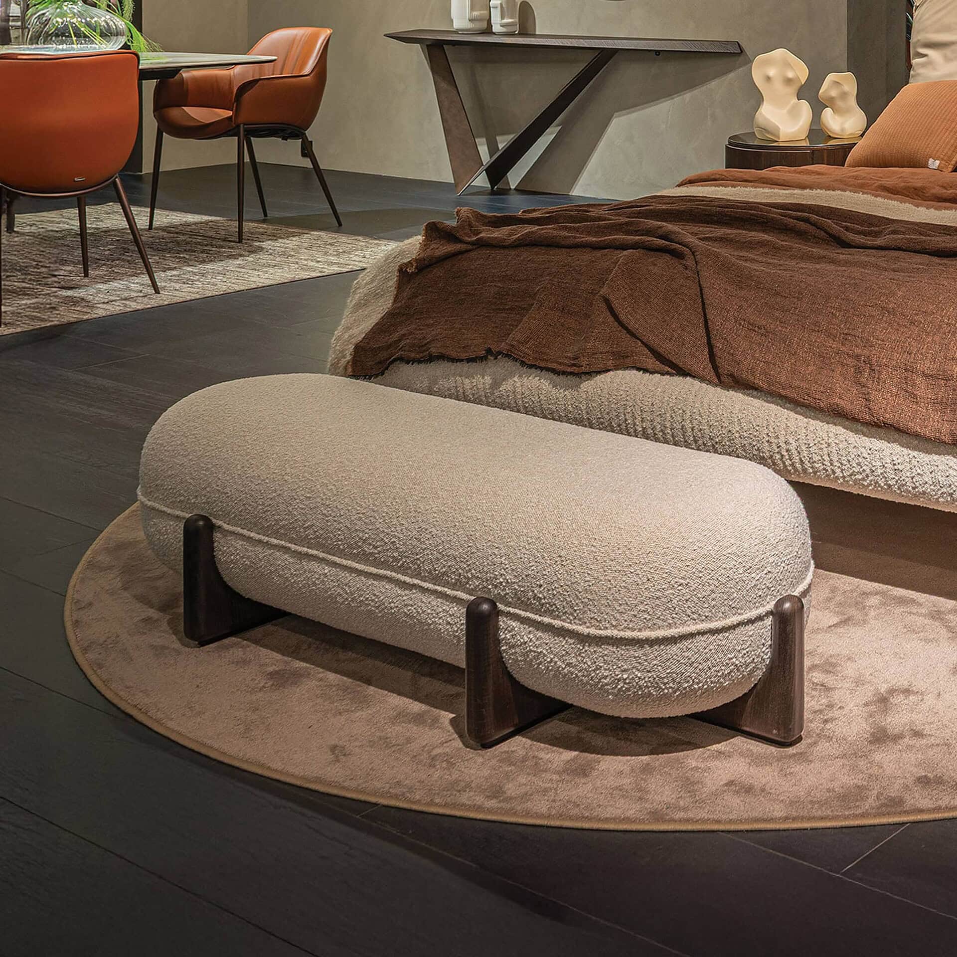 A modern interior with a light boucle bench on a round rug, a bed with brown bedding, and burnt orange leather chairs. A dark console table is in the background.