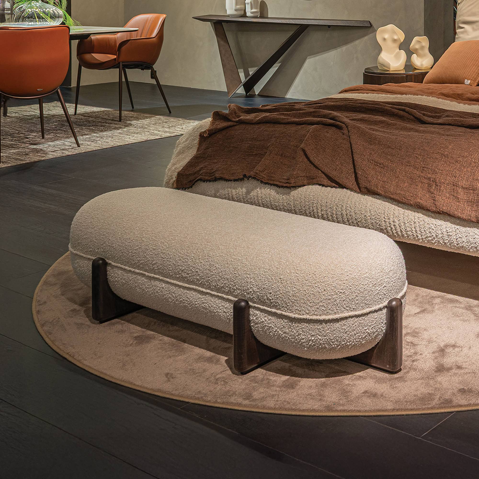 A modern interior with a light boucle bench on a round rug, a bed with brown bedding, and burnt orange leather chairs. A dark console table is in the background.