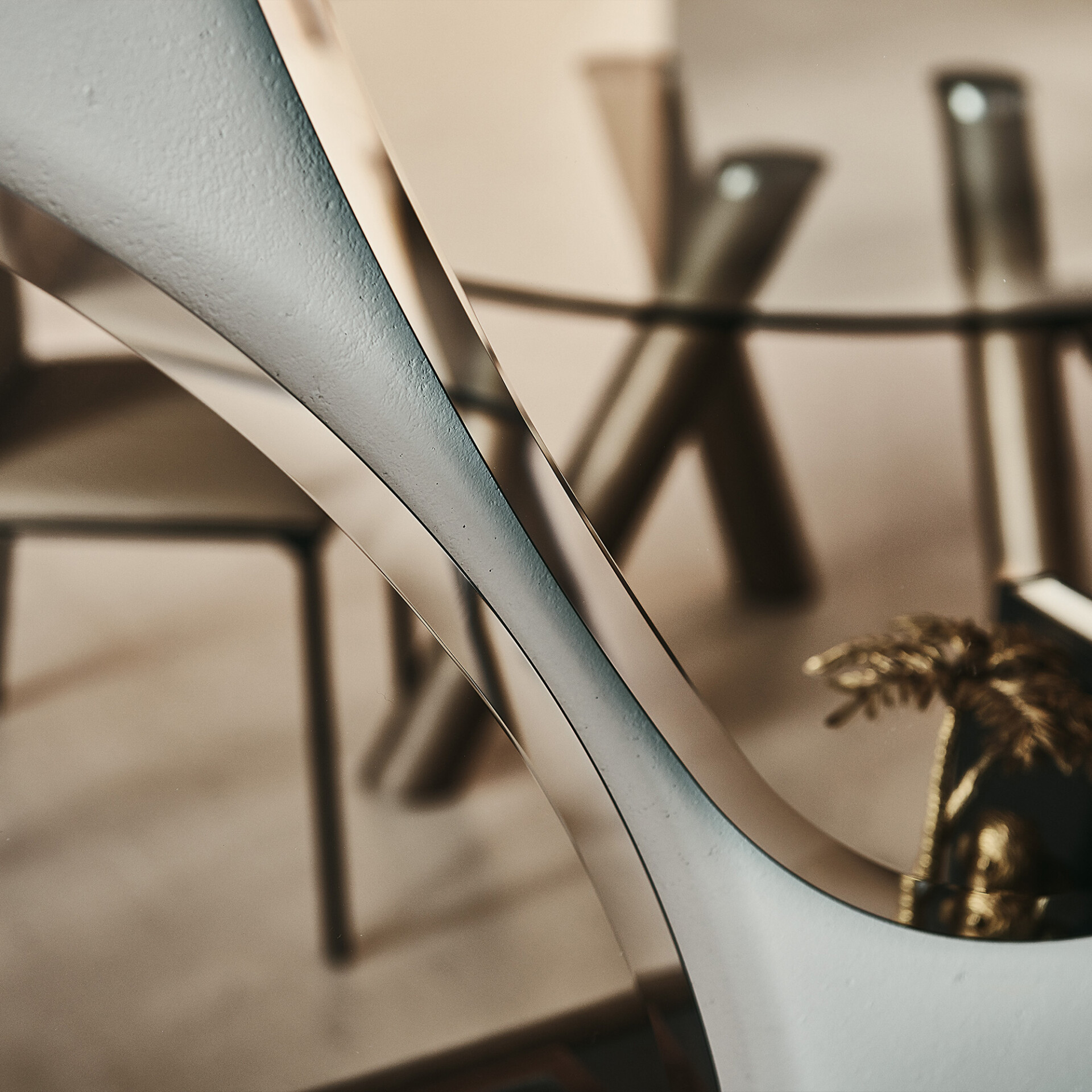 Close-up of a modern decorative object with matte gray and reflective bronze curves. A blurred interior with a table and chairs is in the background.