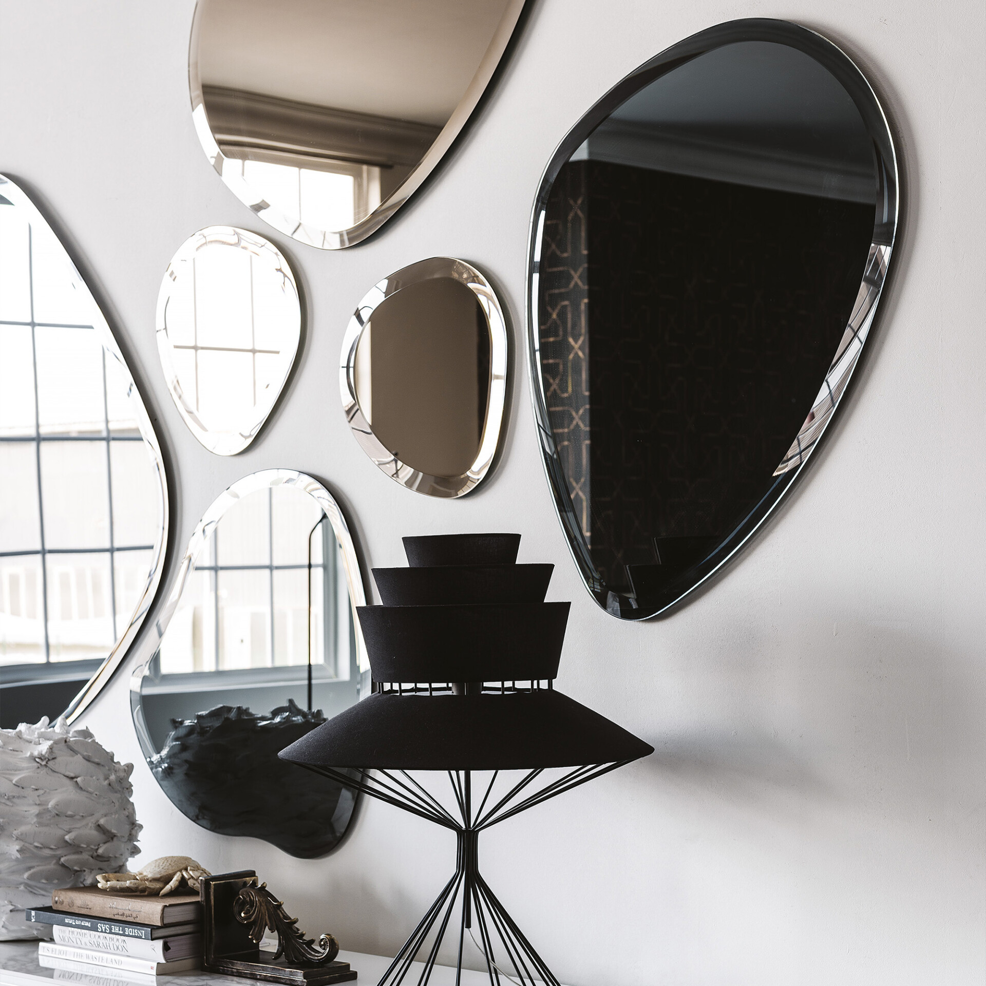 A gallery wall of various irregularly shaped mirrors on a light wall, reflecting windows and a patterned wall. Below, a black sculptural lamp, a stack of books, a decorative crab, and a textured white object are arranged on a console table.
