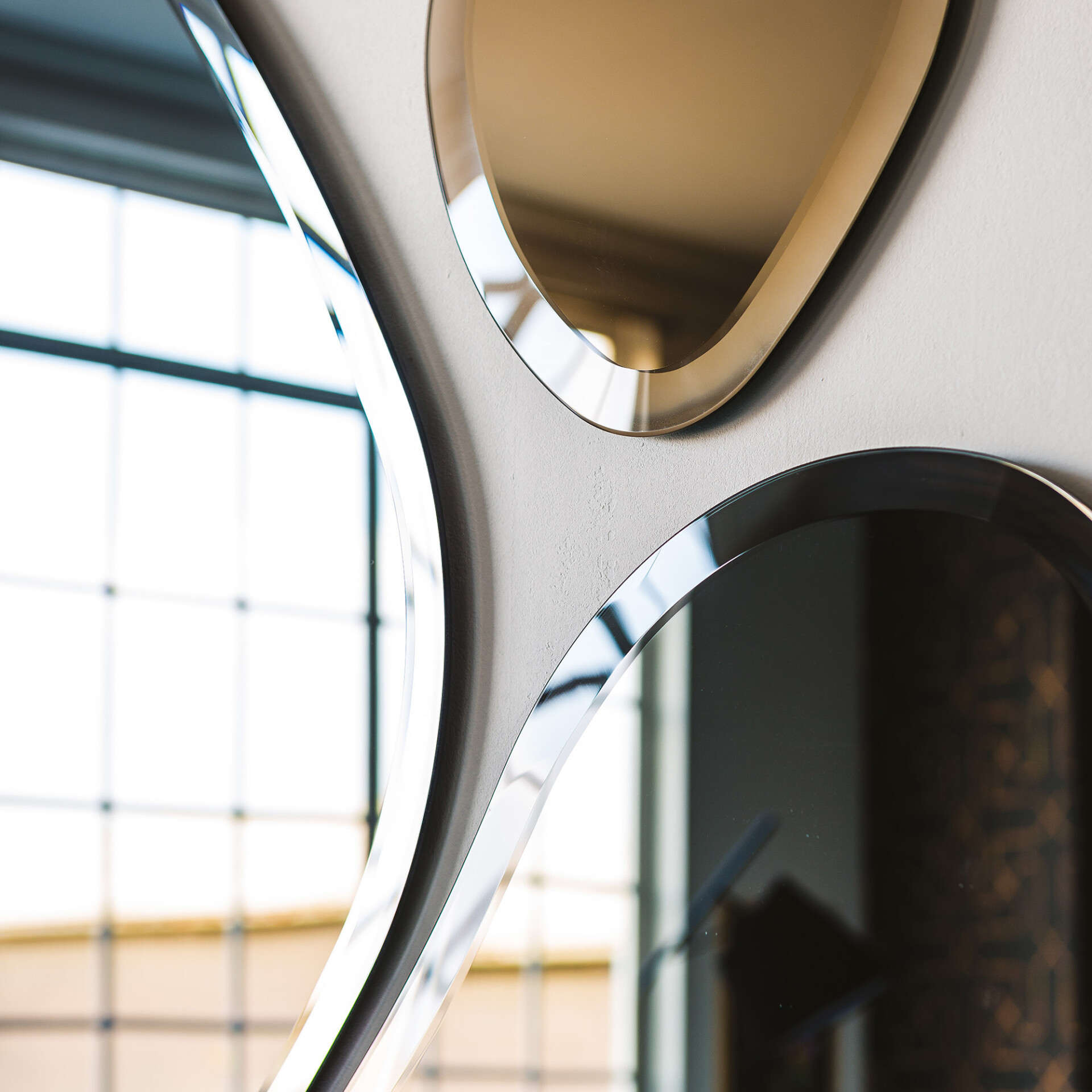 Close-up of a modern wall mirror with two irregularly shaped, beveled glass sections, one golden-brown and one darker, reflecting a bright, gridded window and room details.