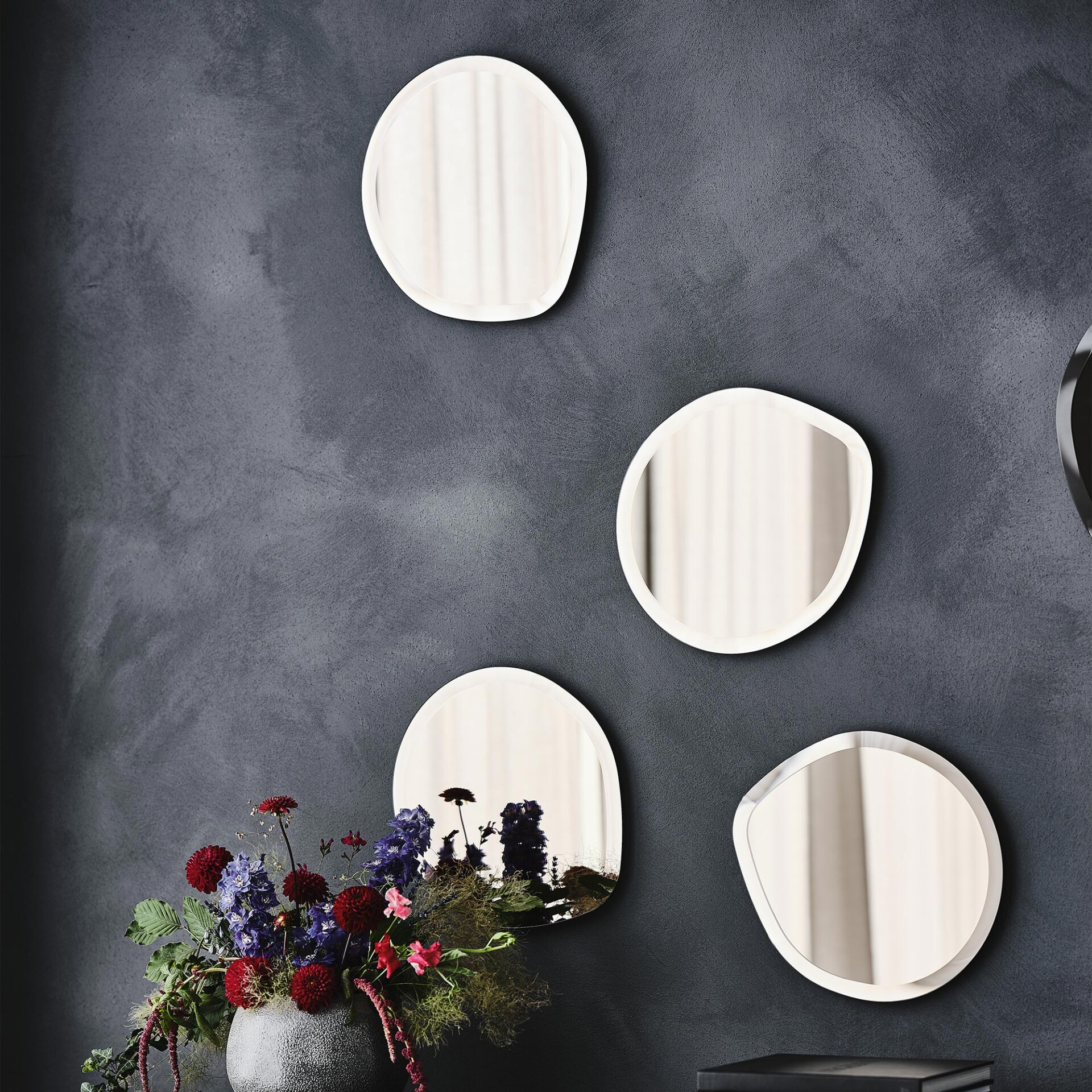 Four irregularly shaped mirrors on a dark, textured wall above a vibrant floral arrangement in a dark vase, featuring red, purple, and pink flowers with green foliage.