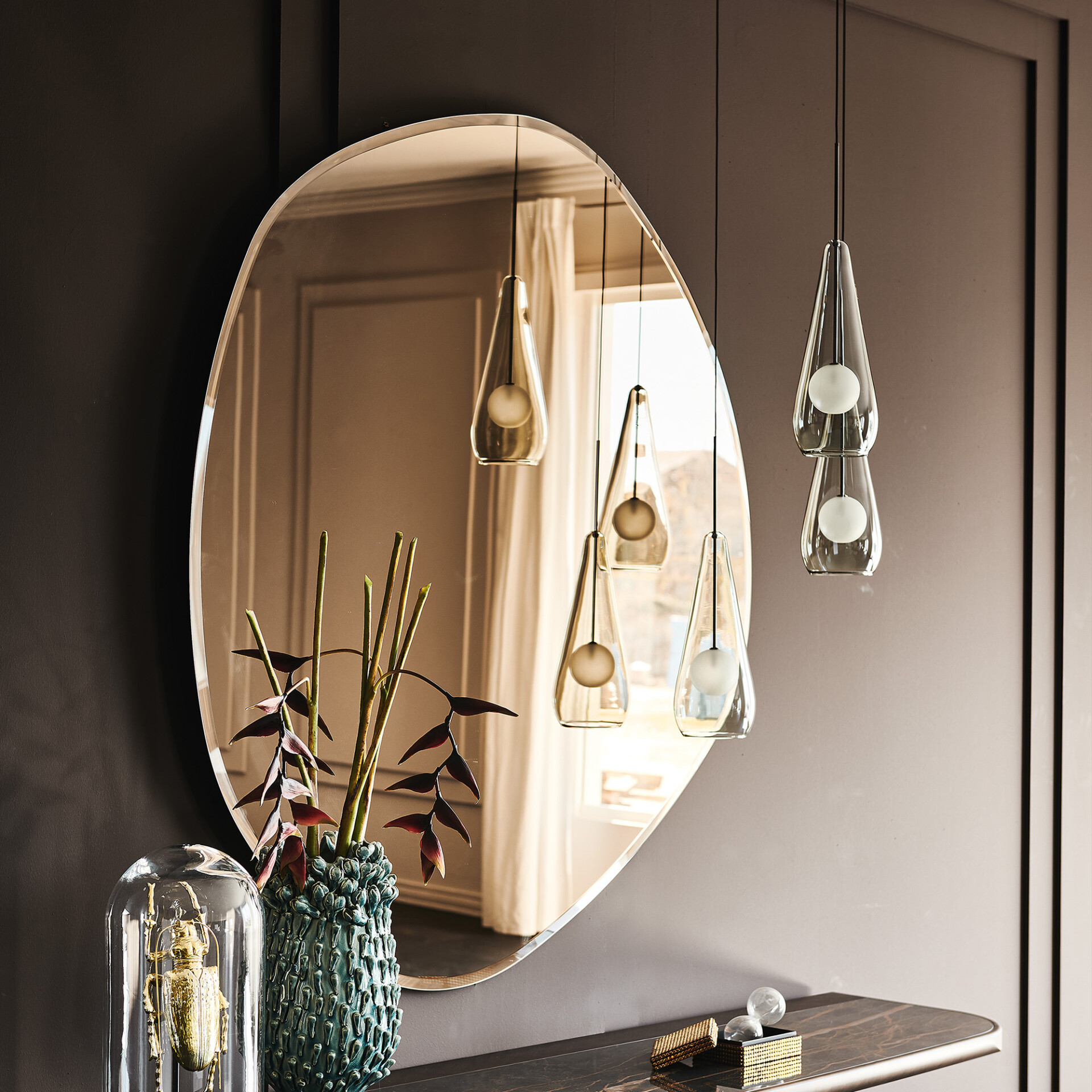 A modern console table setup with an irregular bronze mirror, teardrop pendant lights, a textured teal vase with plants, and a golden beetle sculpture under a cloche.