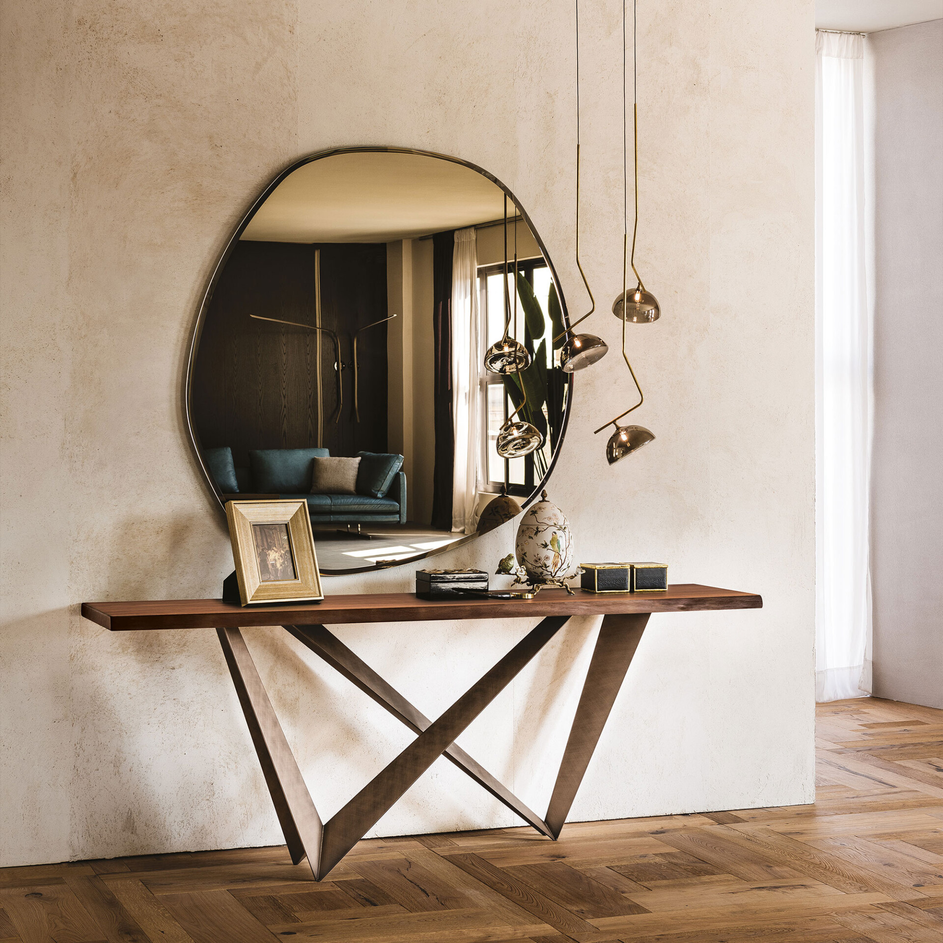 A modern entryway with an oval mirror above a dark wood console table with a geometric metal base. Decorative items, including a gold frame and a ceramic vase, adorn the table. Pendant lights hang to the right, and a herringbone wood floor completes the look.